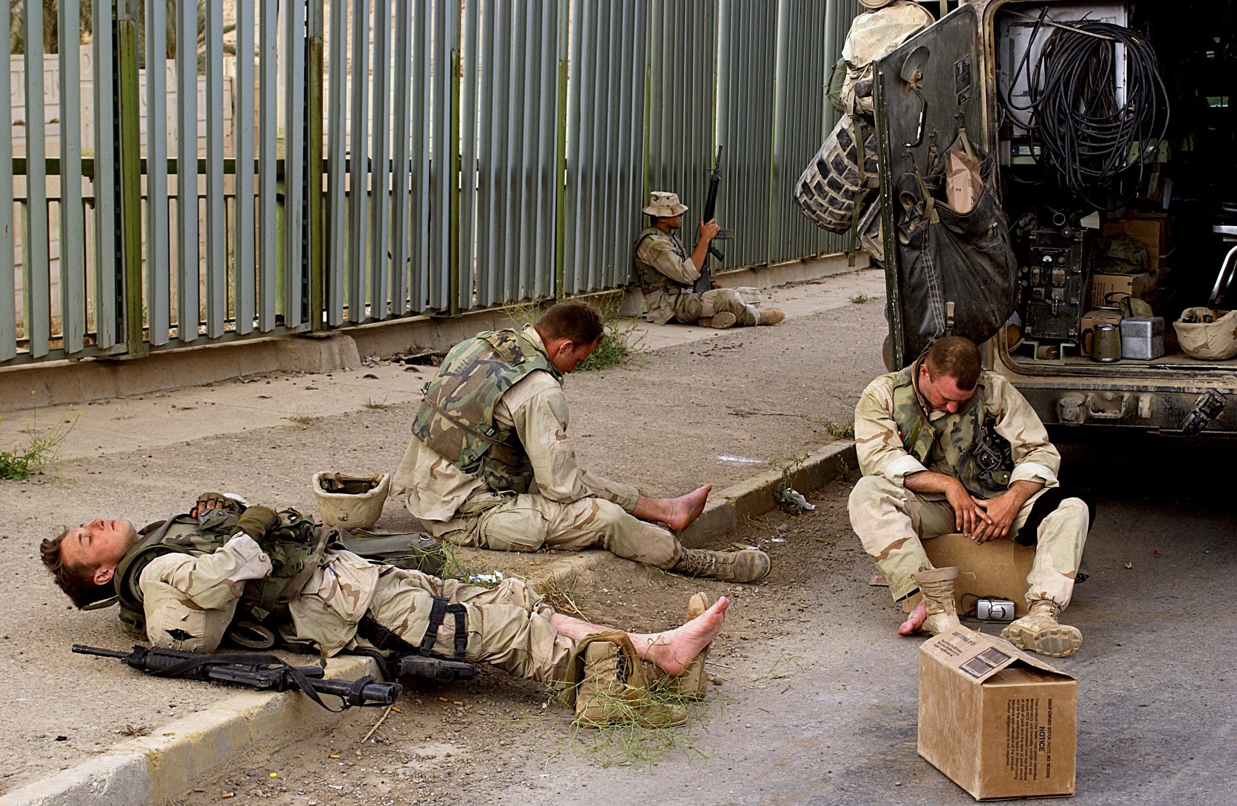 Four soldiers in military uniform resting on a sidewalk near a parked vehicle, with one lying down, another sitting with a prosthetic leg, a third sitting with their head down, and a fourth sitting on the ground near a cardboard box, with a green metal fence in the background.