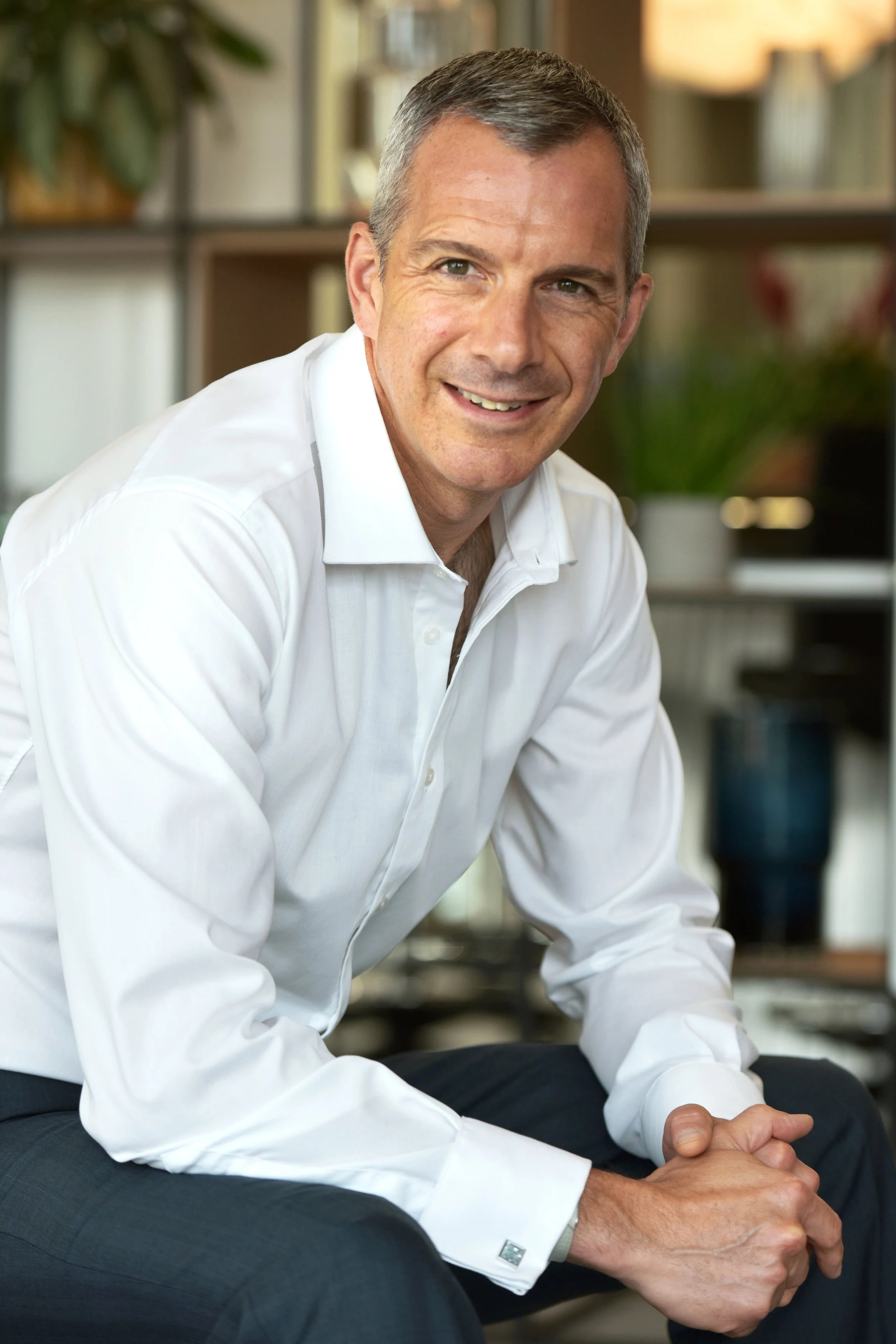 A smiling middle-aged man with short gray hair, wearing a white dress shirt and dark pants, sitting in a modern office with shelves and plants in the background.