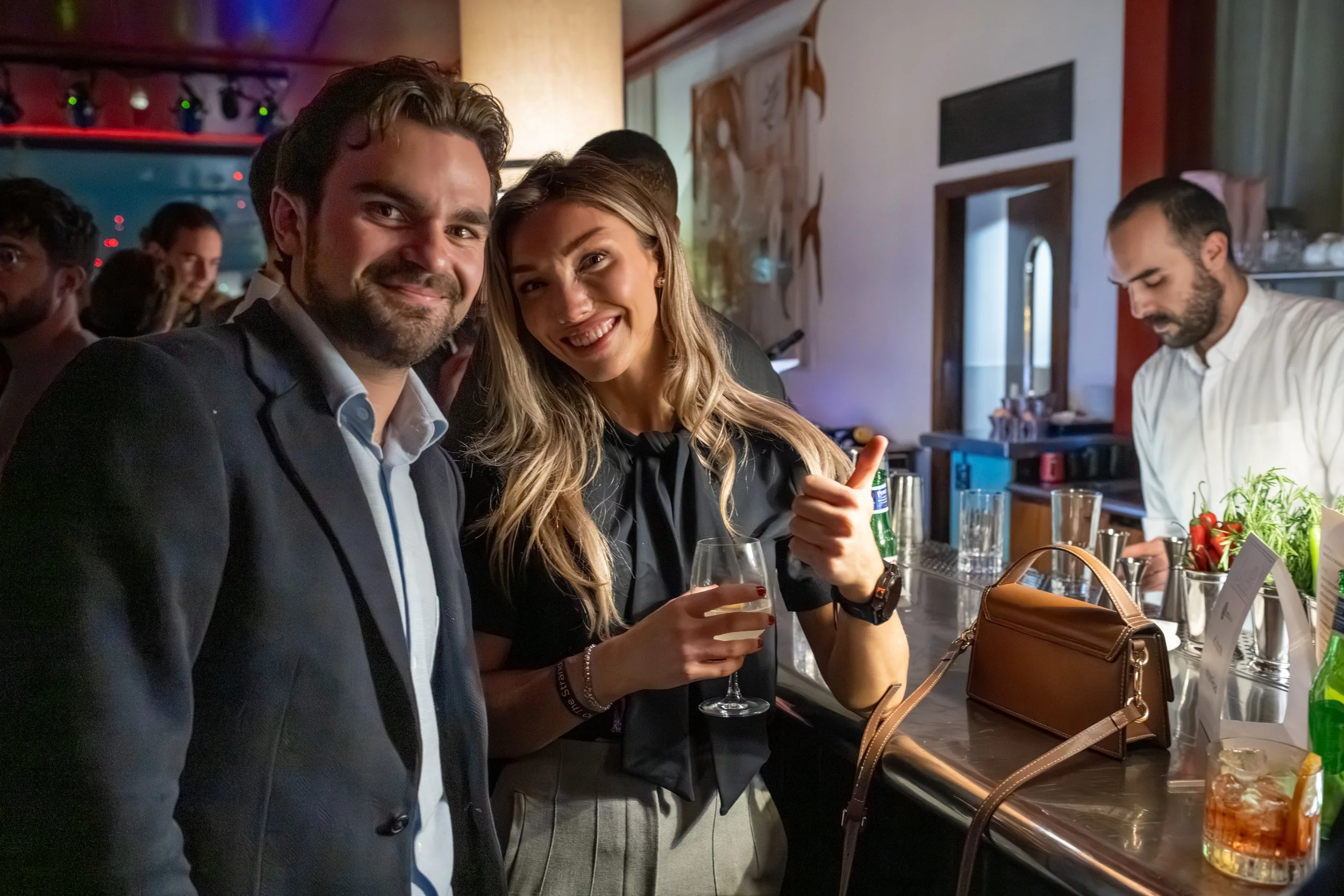 People celebrating at a bar with drinks, with a man and woman in the foreground smiling, and a bartender preparing a drink in the background.