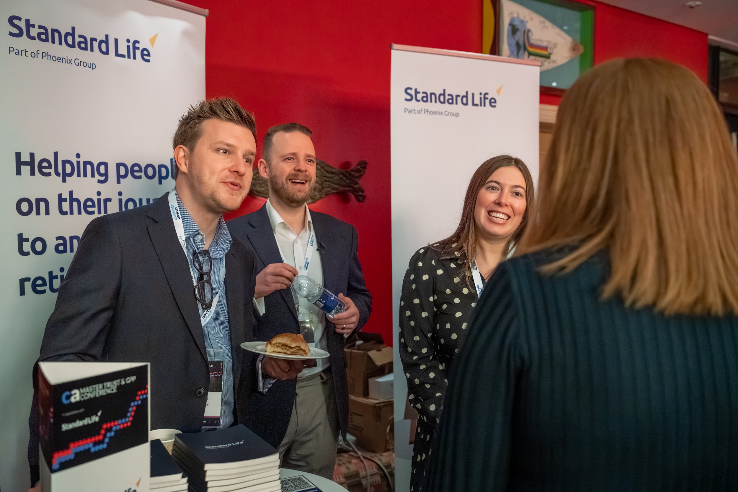 Four people greeting each other at a business event, two men and two women (one from behind), with banners in the background with Standard Life logos and a woman in the foreground, some pamphlets on a table in front.