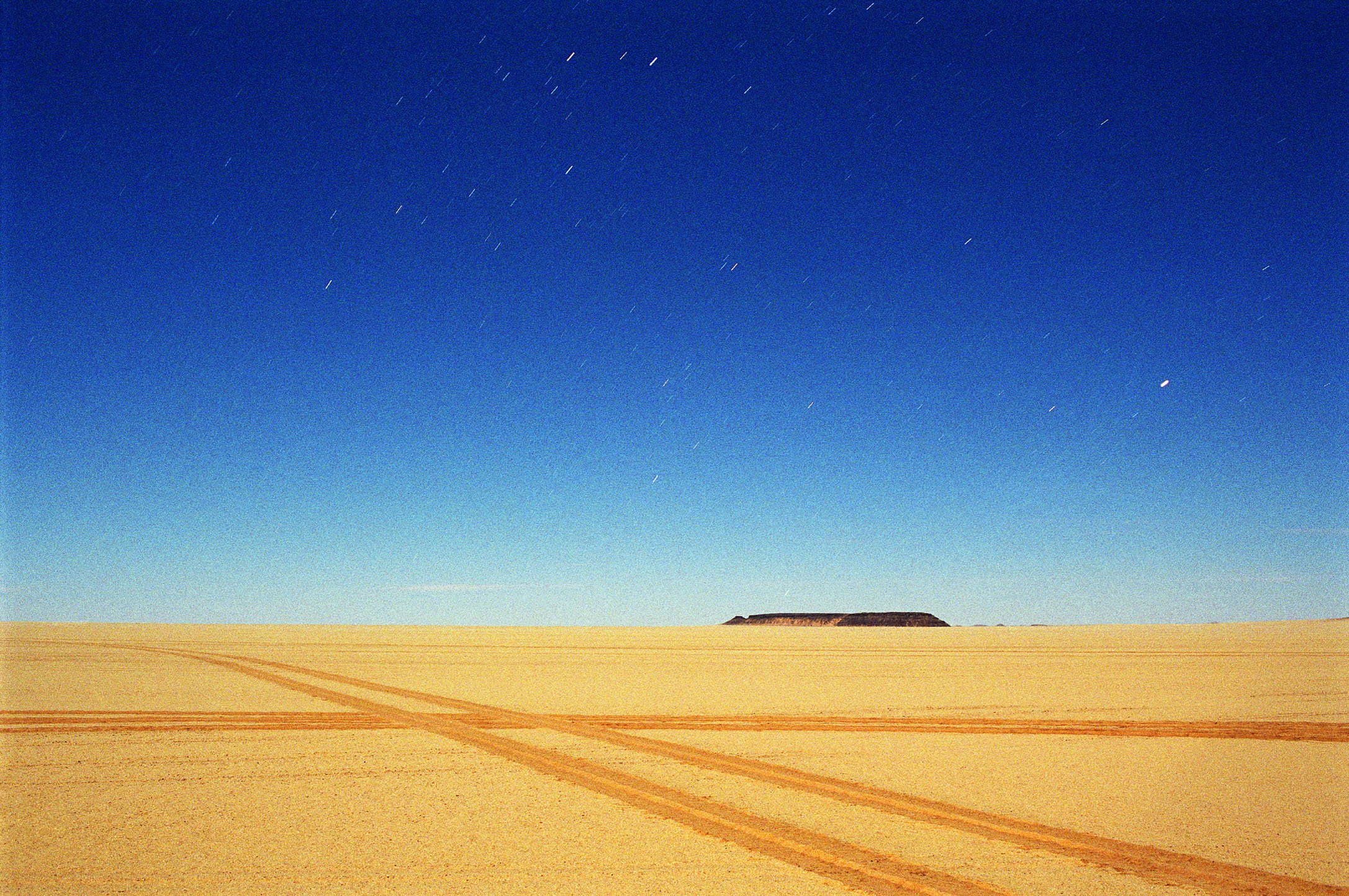 A long exposure night time desert landscape with tire tracks in the sand, a flat-topped hill on the horizon, and a clear blue sky with faint star trails.