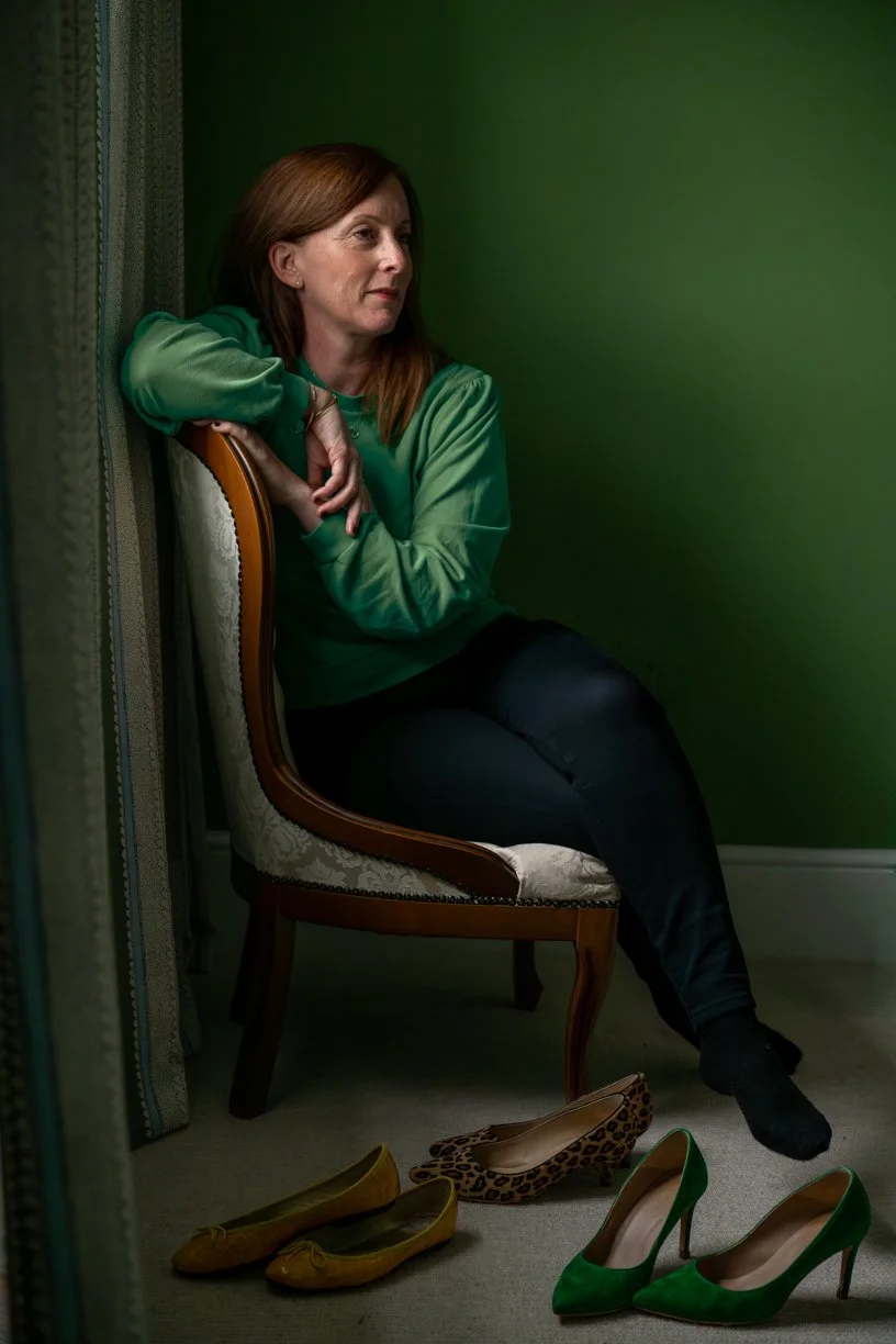 A woman with brown hair sitting on a decorated chair with her arm resting on the back. She is wearing a green satin blouse and black pants. Four pairs of shoes are on the floor in front of her, including yellow flats, leopard print flats, and green high heels. The room has a dark green wall and patterned curtains.