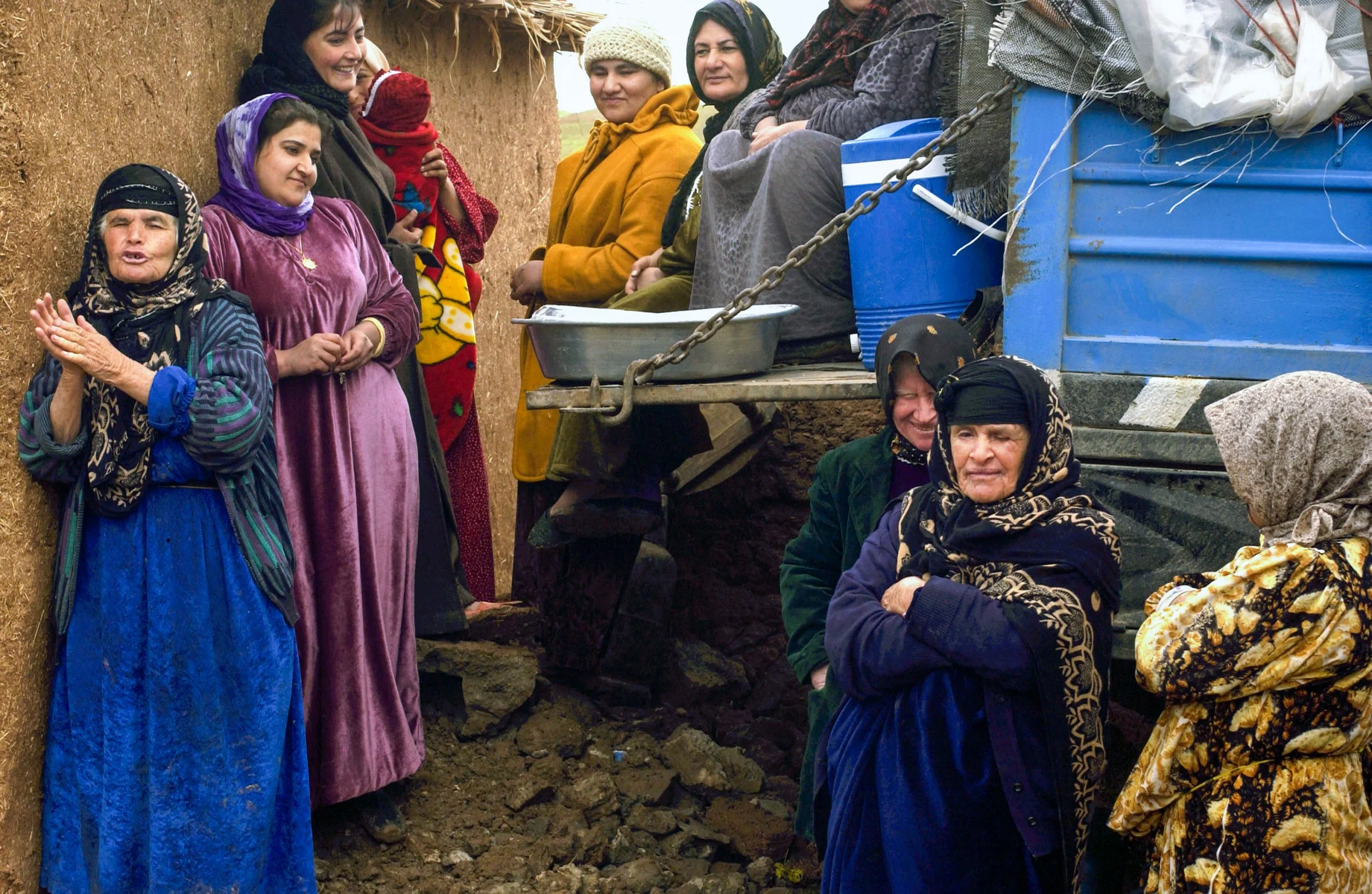 Group of women, some wearing headscarves, gathered outdoors next to a blue truck, engaged in conversation.