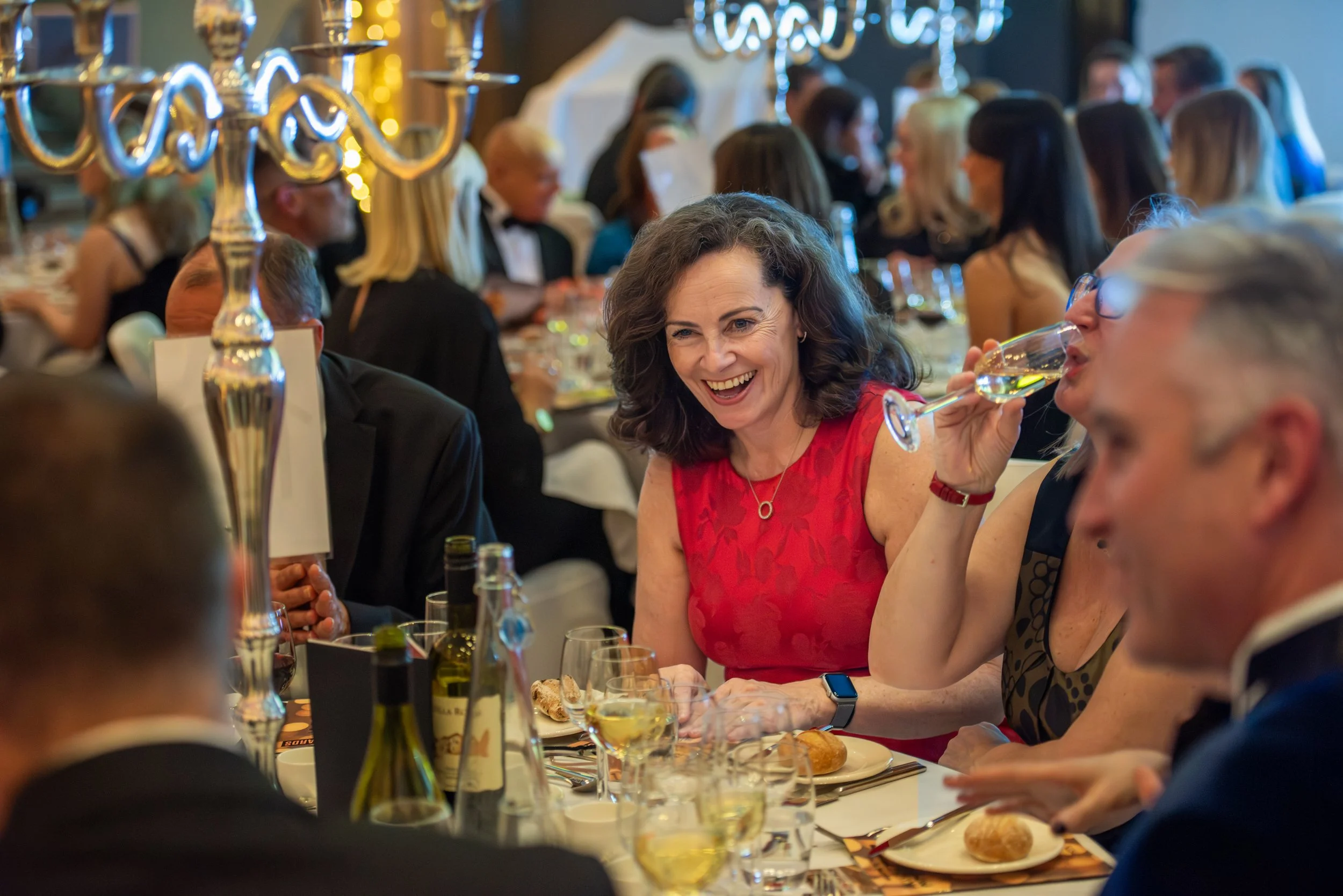 People celebrating at a formal dinner event with a woman in a red dress smiling and holding a glass of wine, surrounded by others at a banquet table with wine bottles, glasses, bread, and decorative candles.