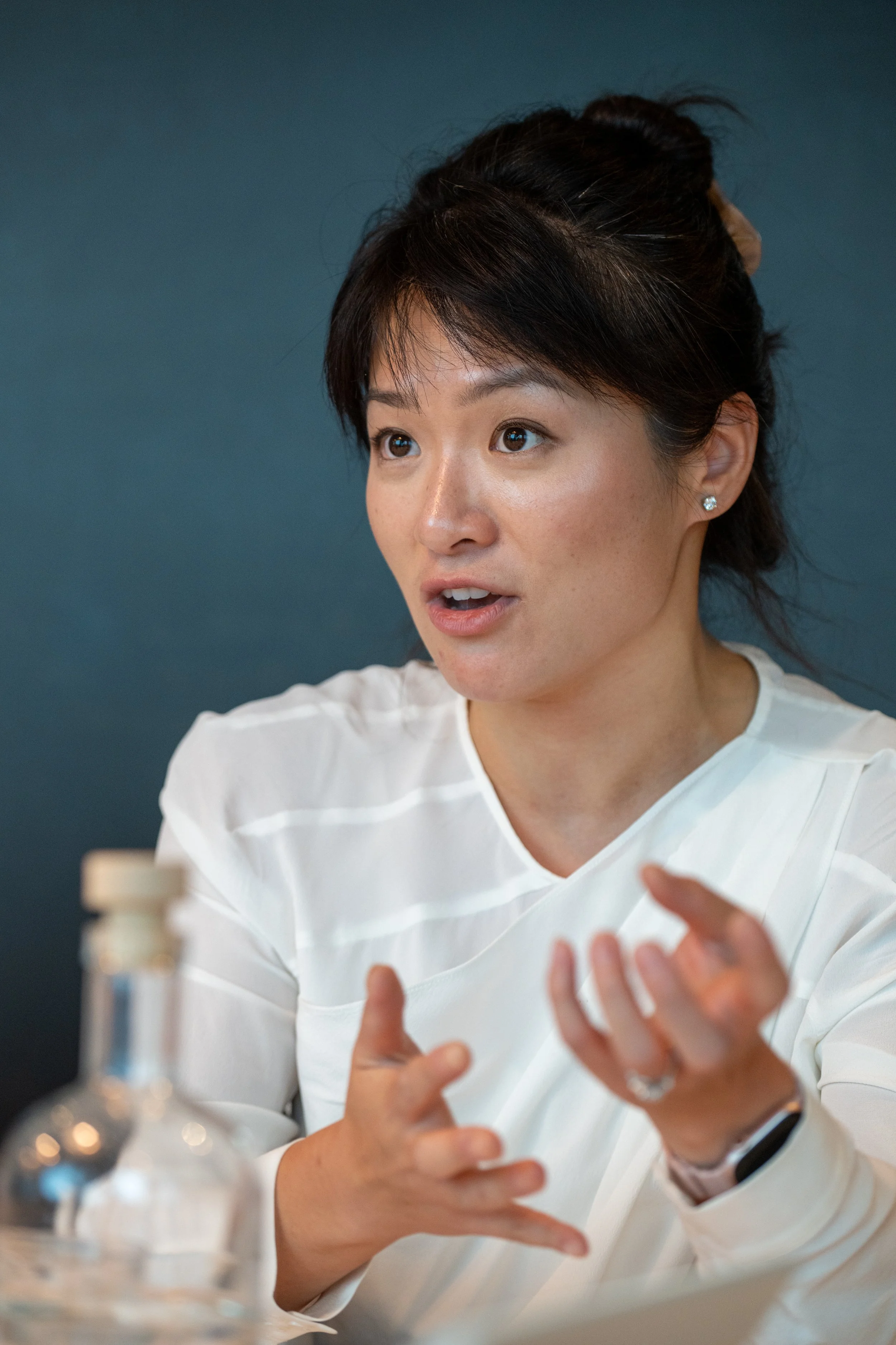 A woman with dark hair tied back, wearing a white blouse, appears to be engaged in a conversation or discussion, with a serious expression on her face and her hands gesturing.