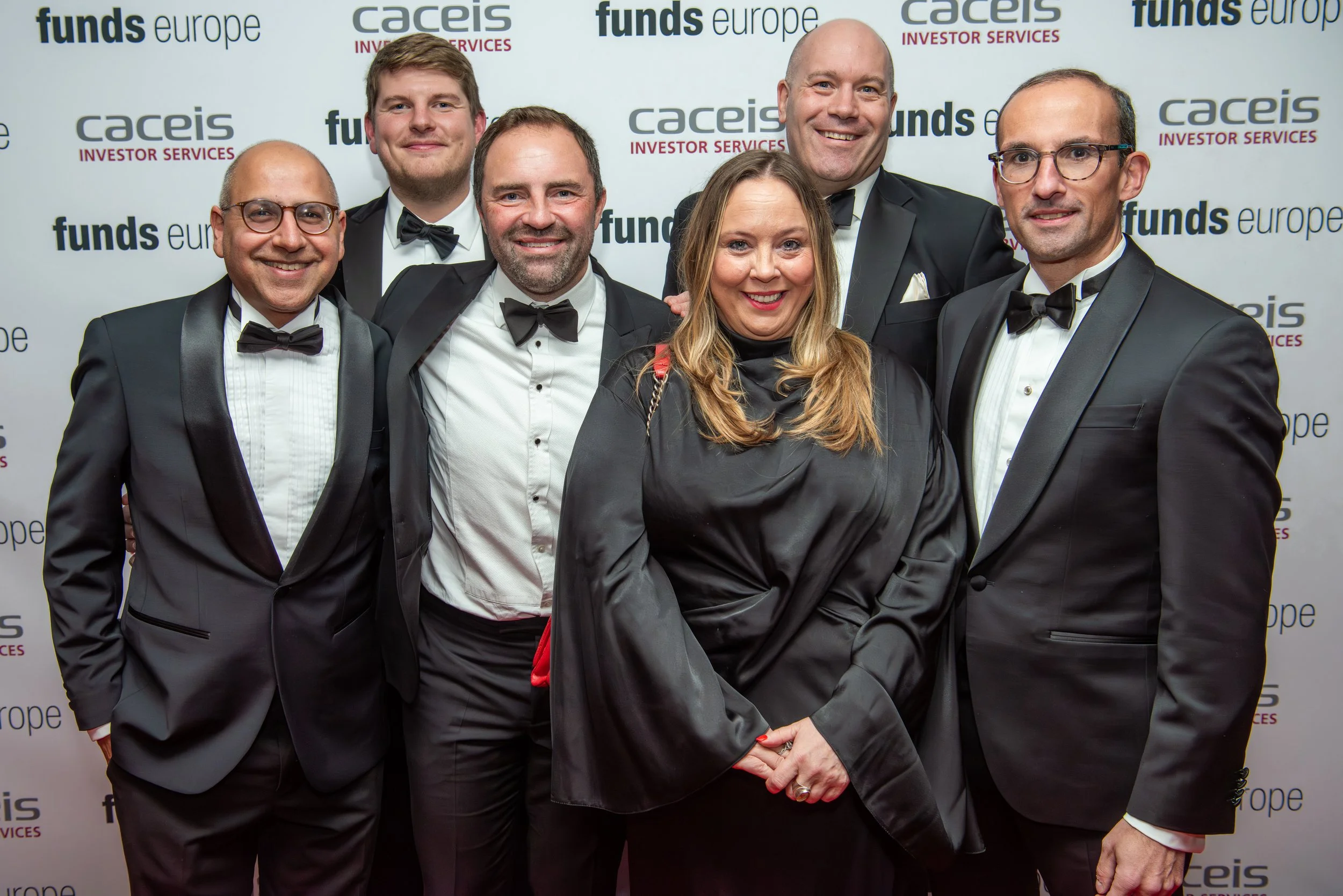 A group of seven diverse people dressed in formal black-tie attire, posing for a photo at a professional event with a white backdrop featuring logos for 'Caceis' and 'Funds Europe'.