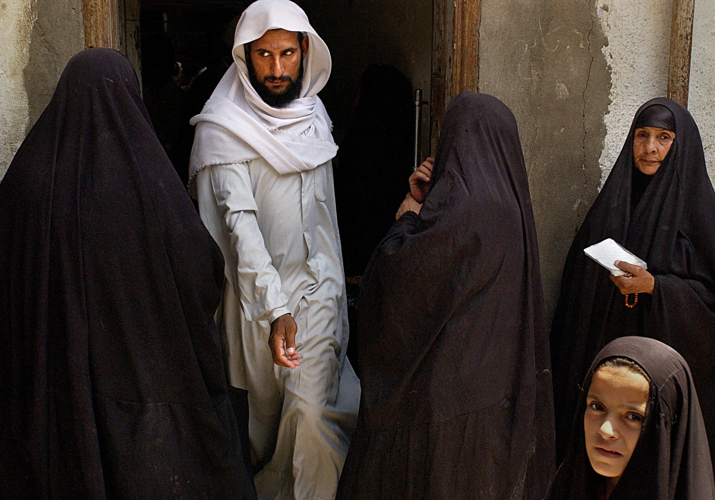 A group of five people, four women in black hijabs and one man in white traditional clothing, standing outside a weathered building entrance with a wooden frame.