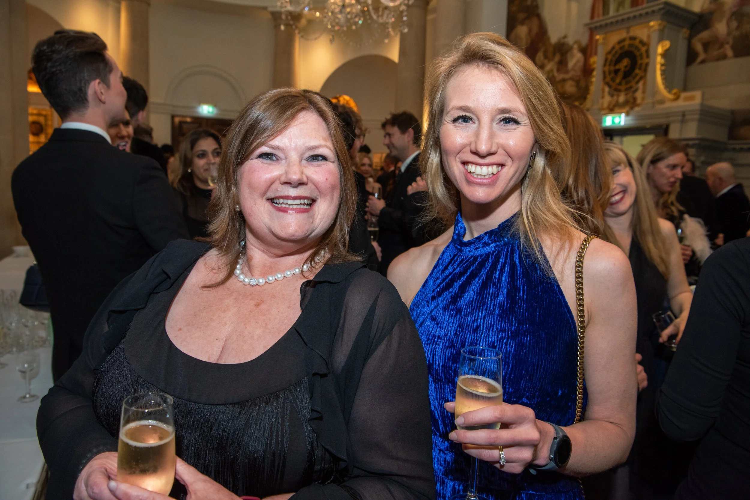 Two women smiling and holding glasses of champagne at a formal event, with a crowd of people in the background.