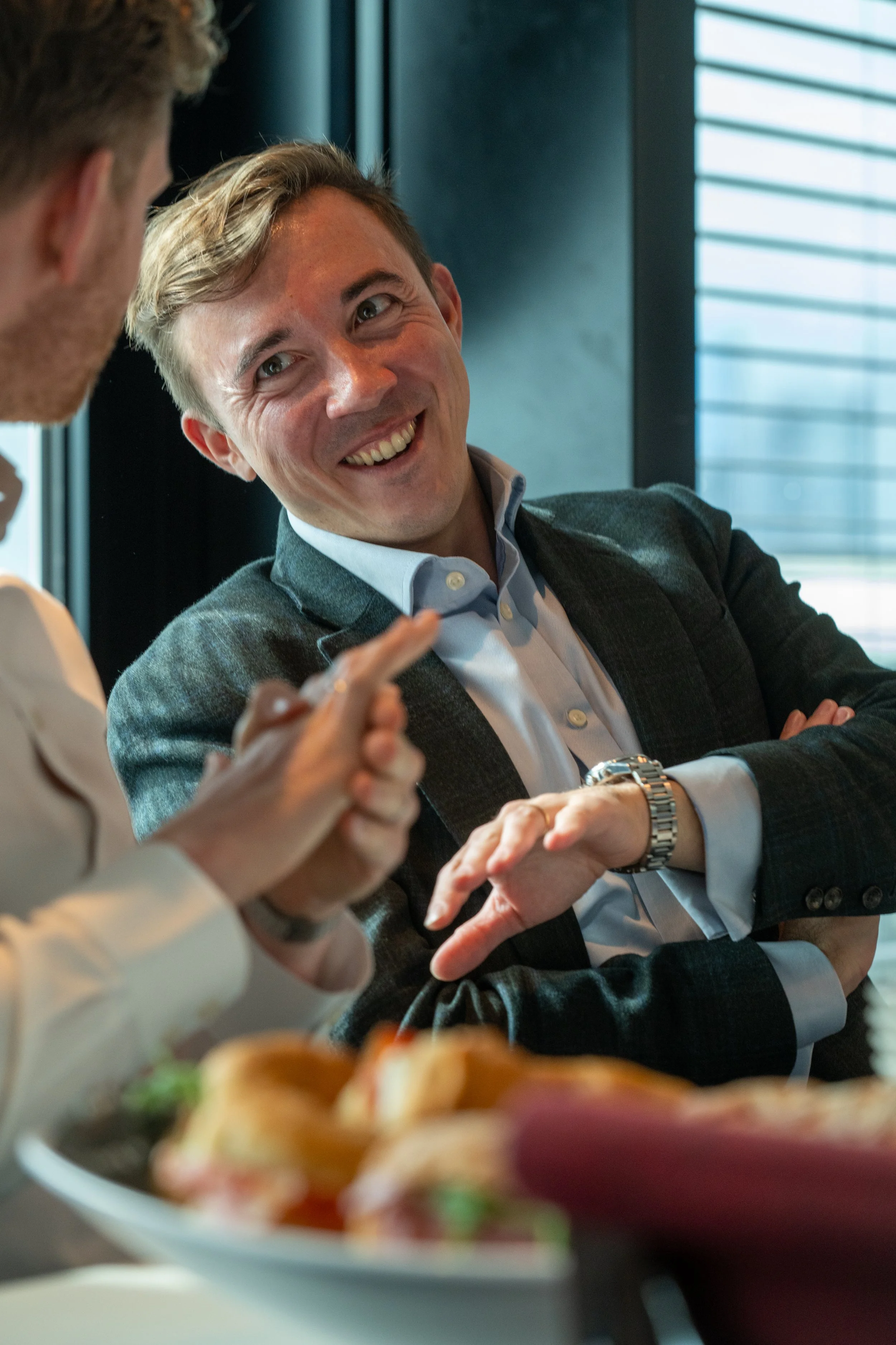 Two men are having a conversation at a restaurant, with one smiling and the other gesturing. There is a plate of sandwiches in the foreground.