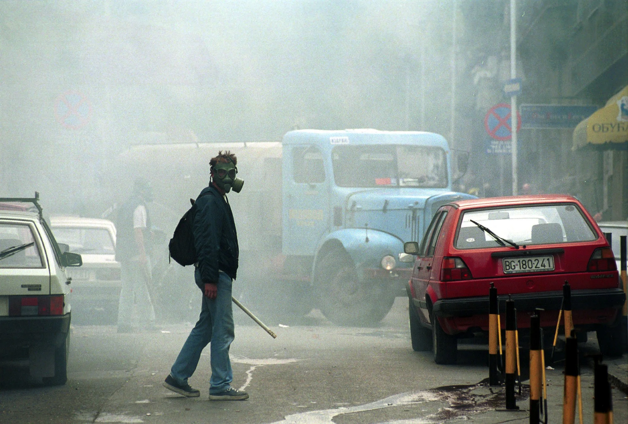 A person wearing a gas mask and backpack walking through a street filled with thick smoke, surrounded by parked cars and old vehicles, with shop signs visible in the background.