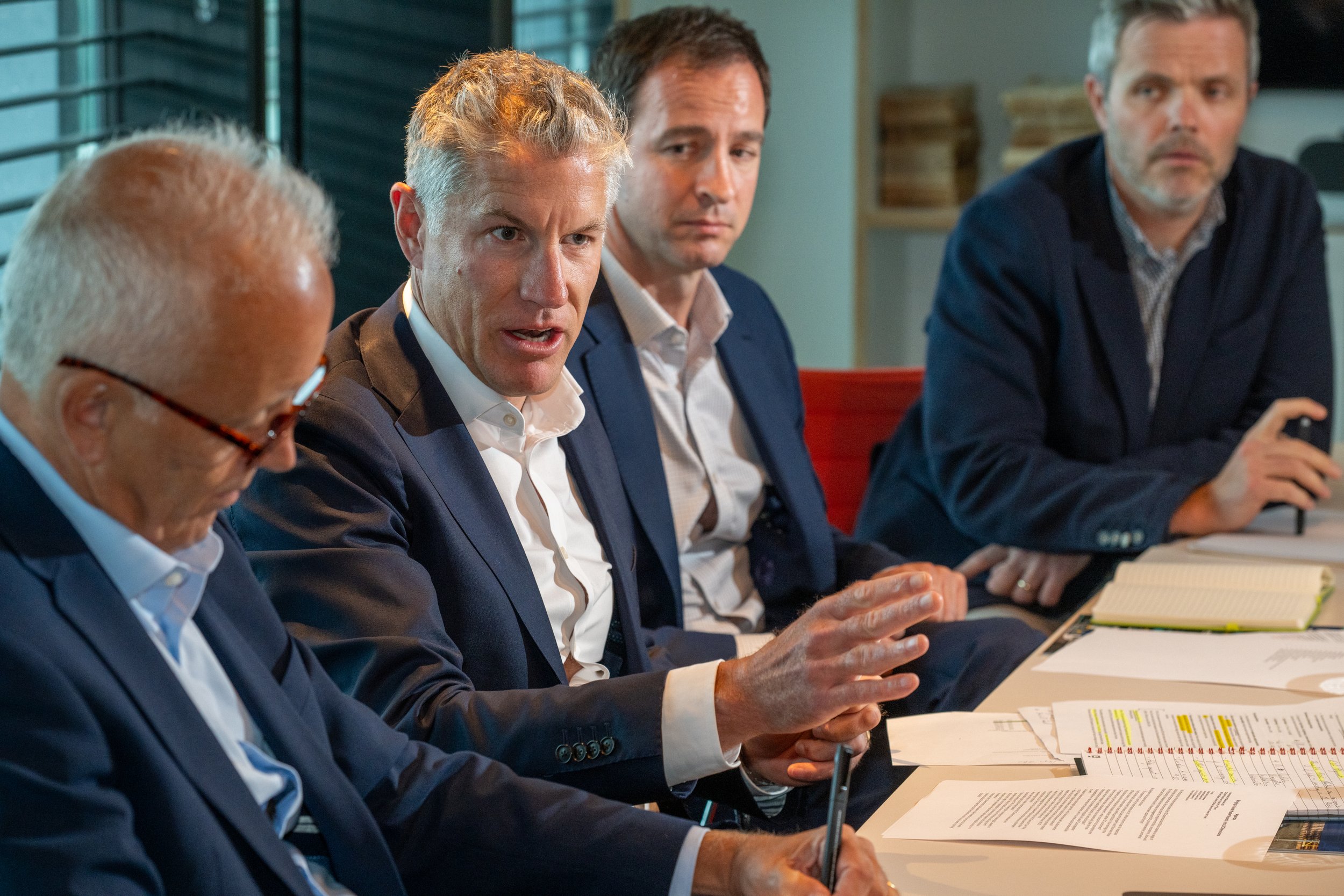 Four men in suits sit around a table during a meeting, with documents and notebooks in front of them.