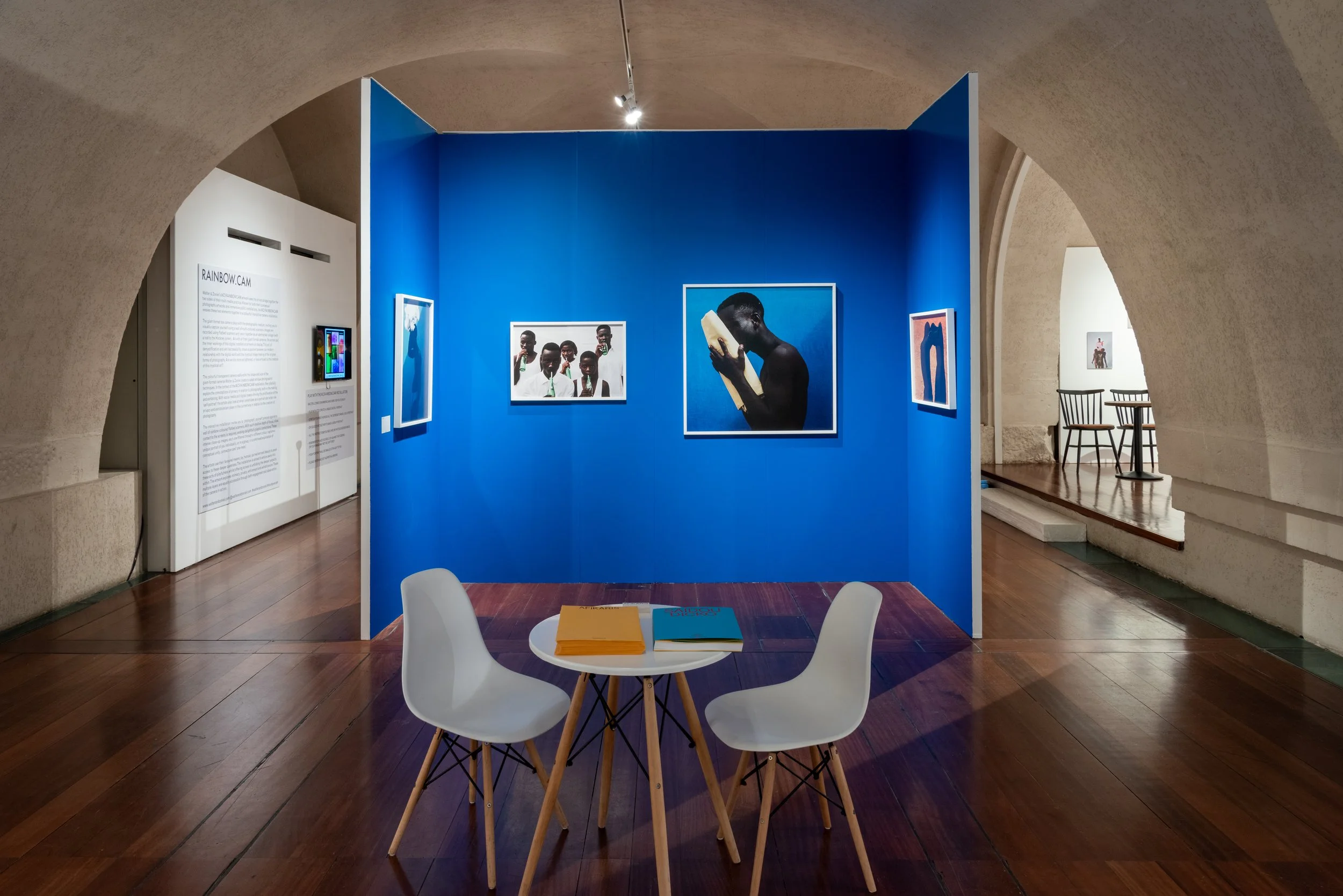 An art gallery with vibrant blue walls displaying photographs. In the foreground, a small round table with two white chairs holds two books. Adjacent walls and a distant sitting area are visible.