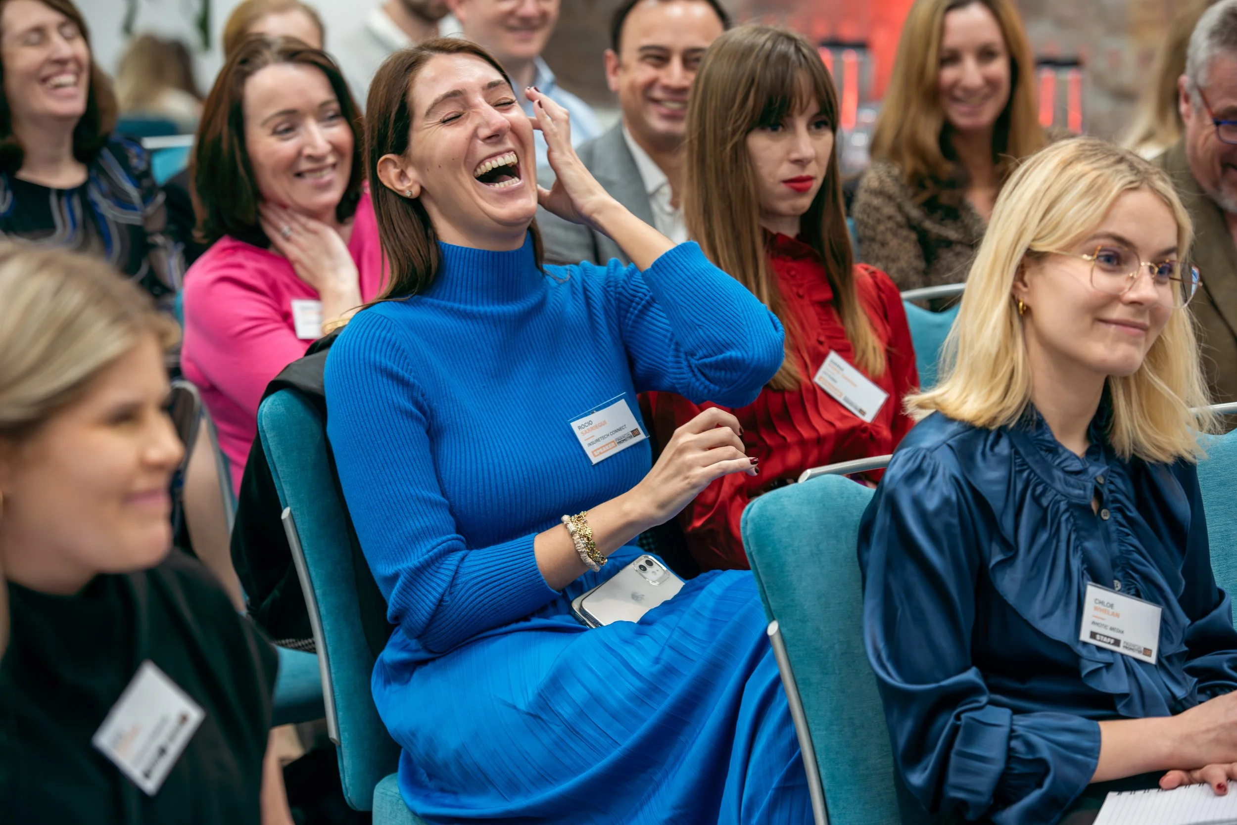 Group of people attending a conference or seminar, enjoying and laughing, seated in chairs in a professional setting.