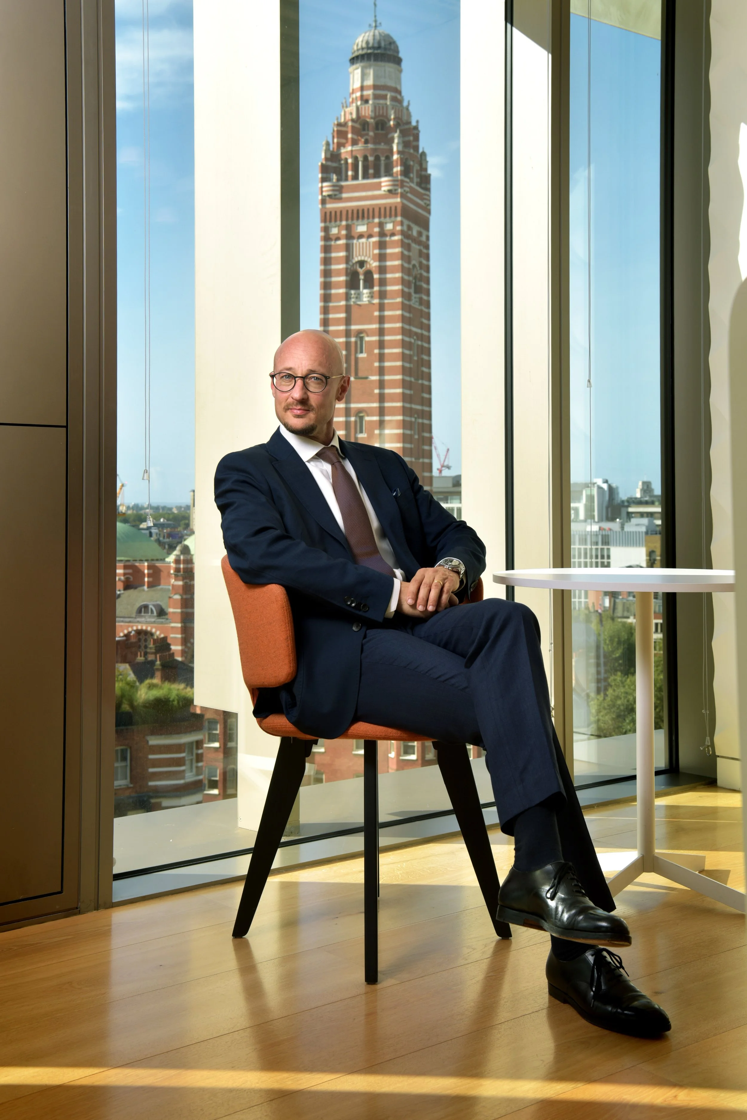 A man in a suit sitting on a chair in an office with large floor-to-ceiling windows, overlooking a cityscape with Westminster Cathedral's brick clock tower in the background.
