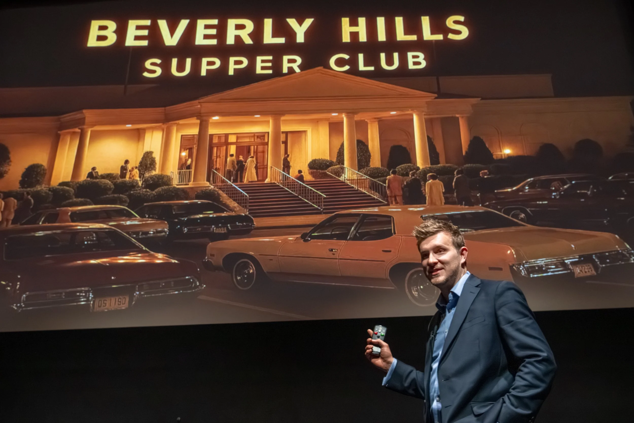 A man in a blue suit holding a remote control standing in front of a large screen displaying an image of the Beverly Hills Supper Club with vintage cars parked outside at night.