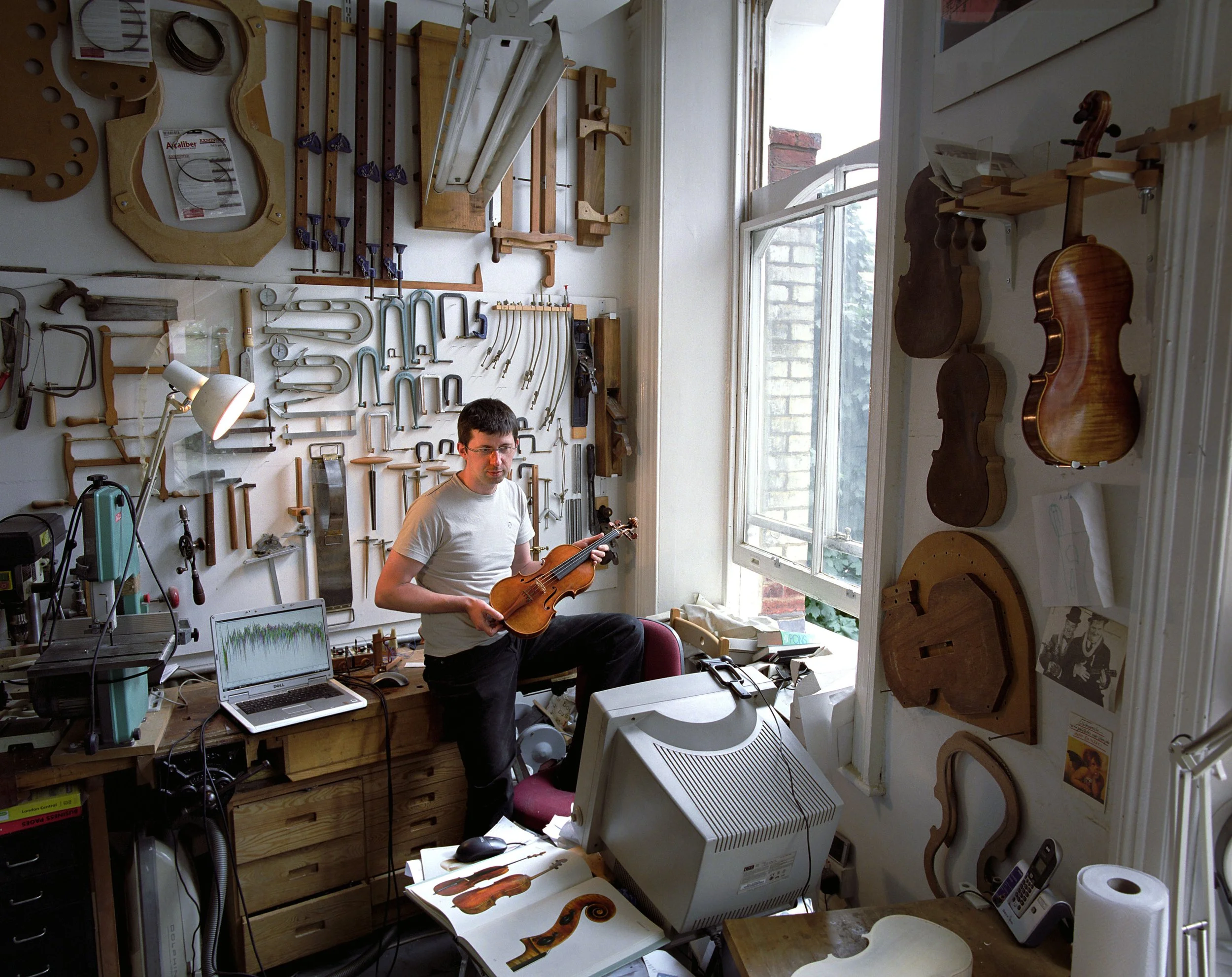 A man in a workshop holding a violin, surrounded by various woodworking tools and partially assembled violins on the wall and table.