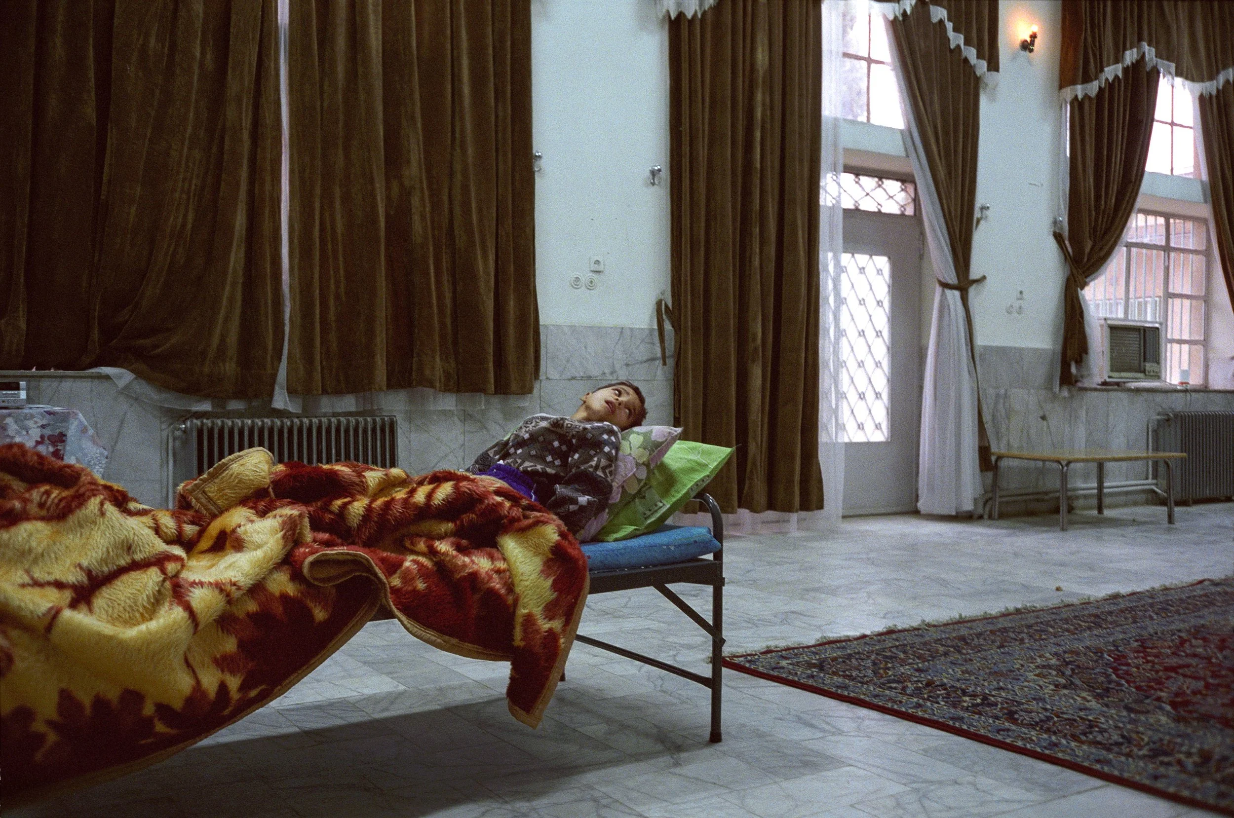 A woman lying on a hospital bed with a colorful blanket in a room with brown curtains, a patterned rug, and large windows with curtains drawn to the sides.