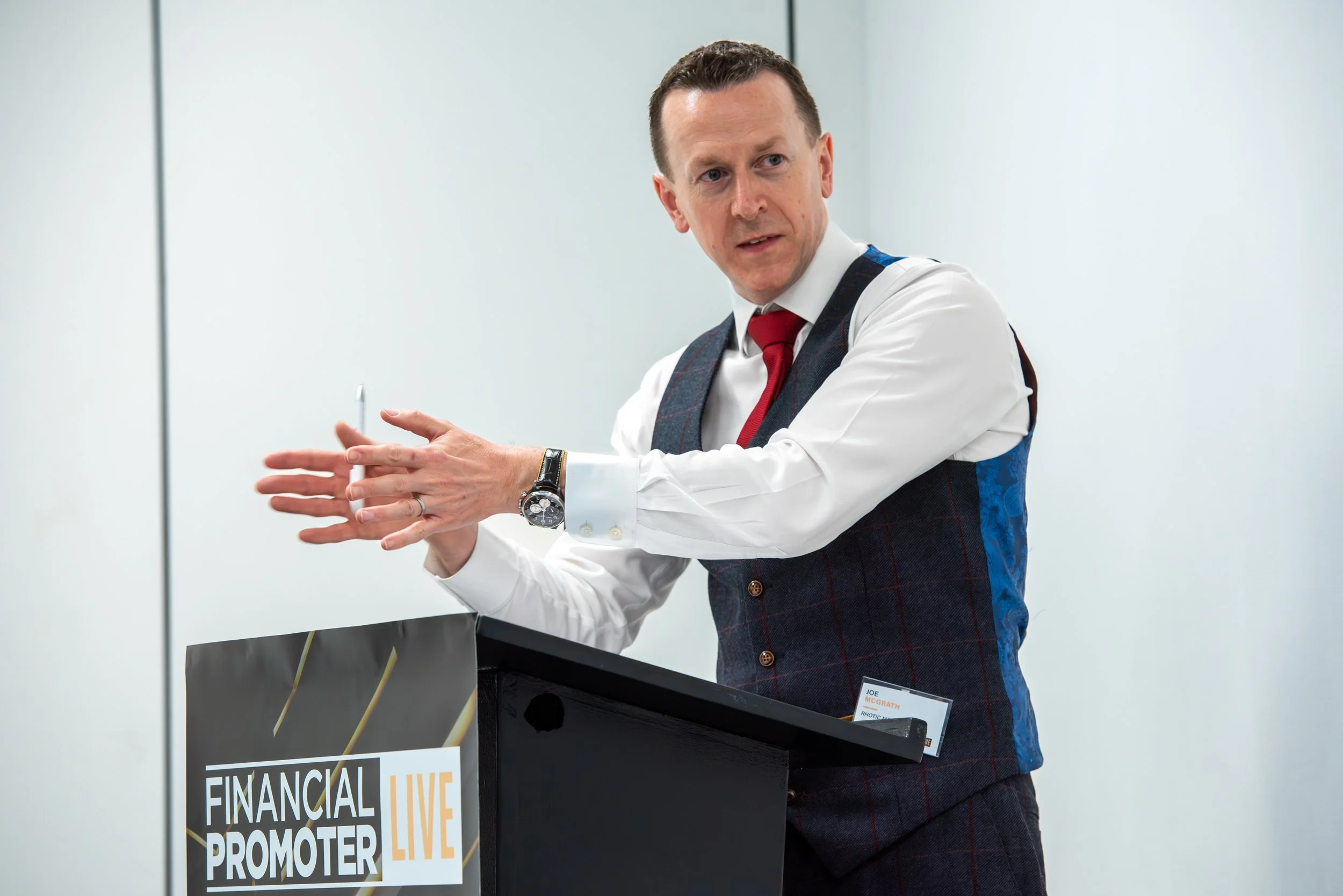 A man in business attire, wearing a white shirt, red tie, and a dark vest, is standing behind a podium labeled 'Financial Promoter Live.' He appears to be speaking or presenting, with one arm extended and a focused expression on his face.