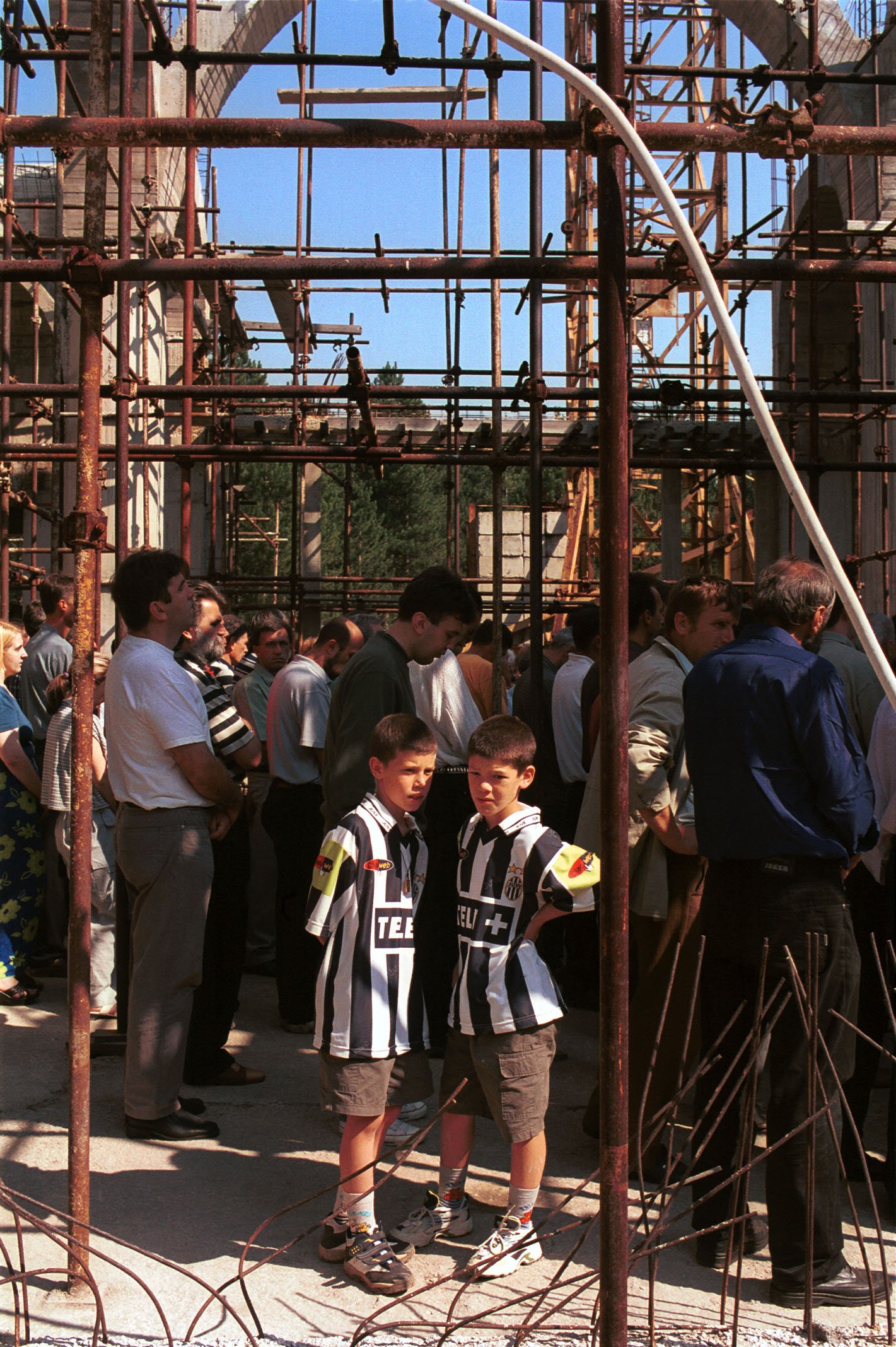 A group of people, including two boys in black and white striped soccer jerseys, standing behind a rusty metal barrier at a construction site or scaffolding structure during daytime.