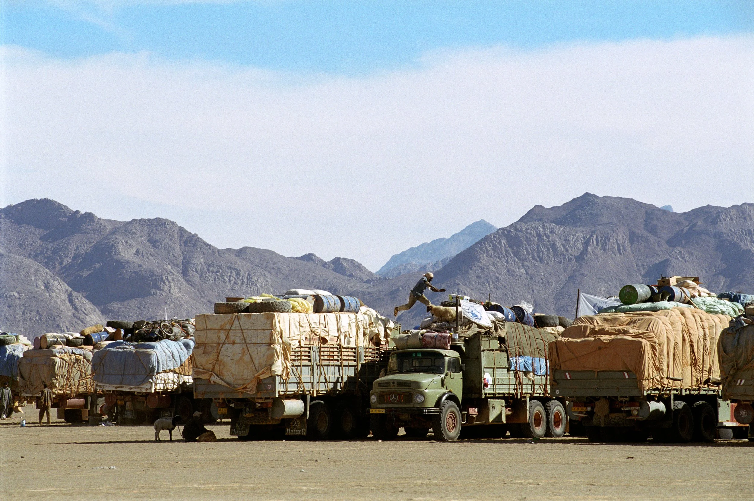 Aid trucks loaded with supplies in a desert with mountains in the background, and a person jumping from one truck to another.