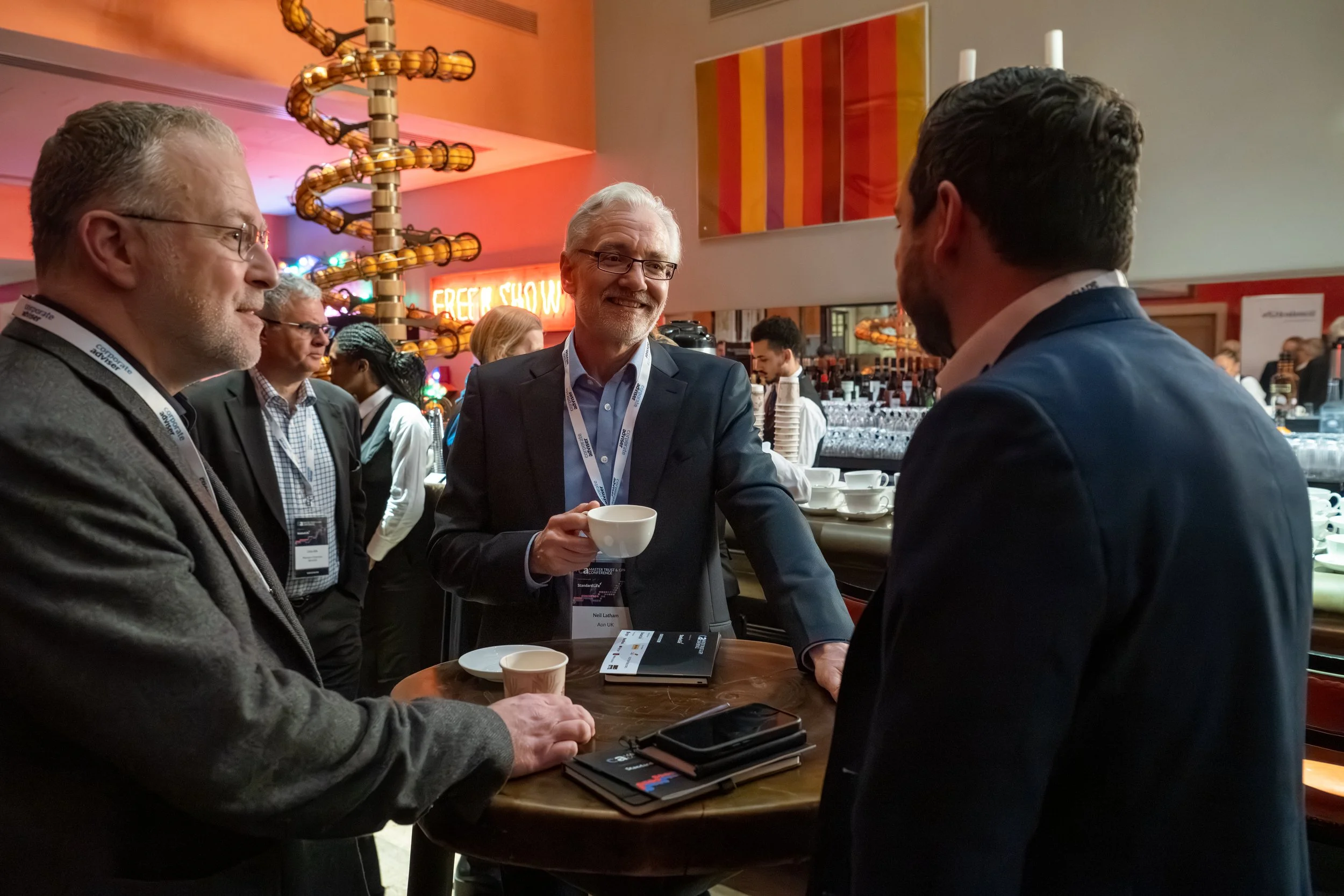Four men in business attire engaged in conversation at a bar during a conference, with a bar setup, colorful decorations, and people in the background.