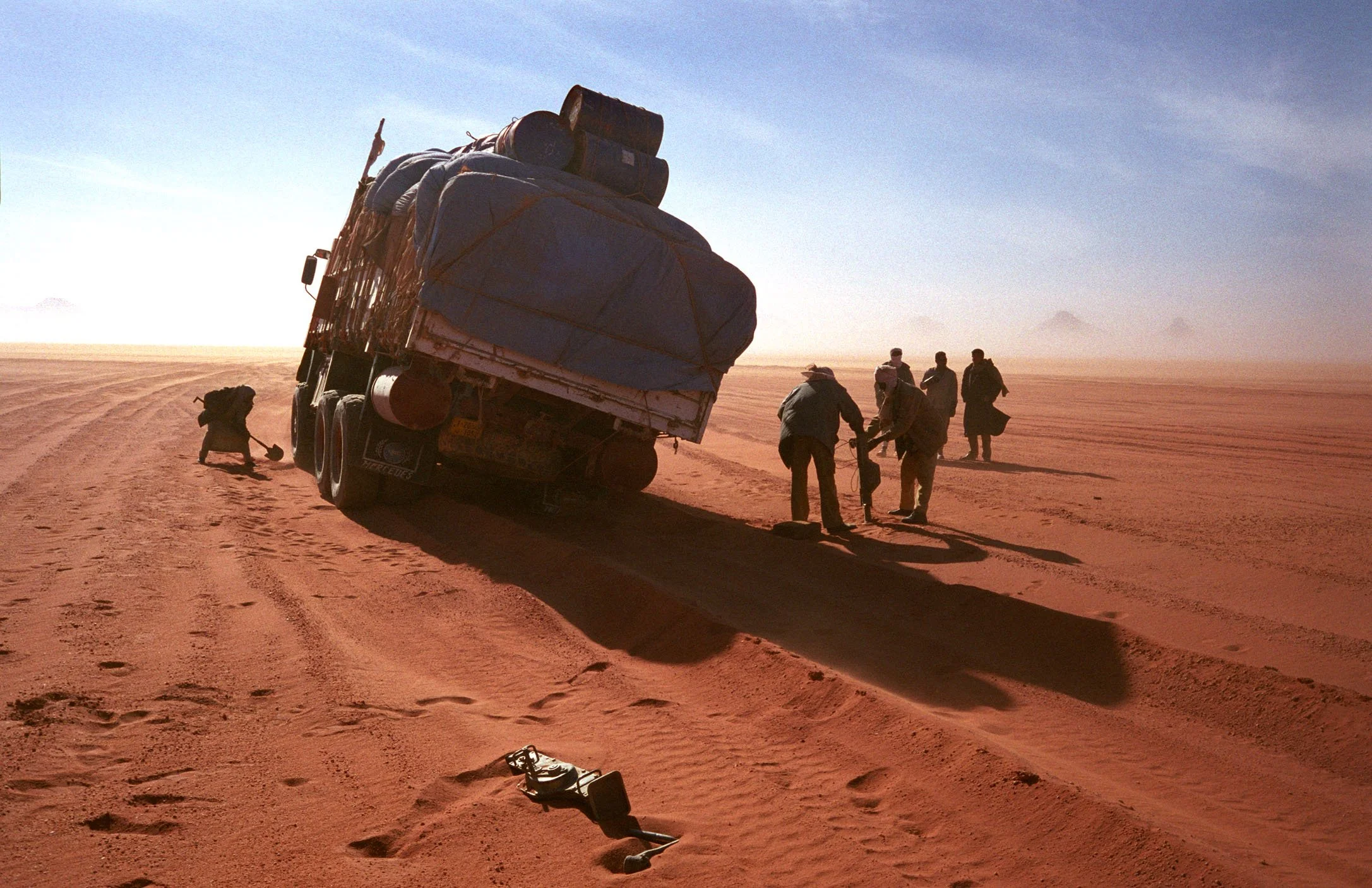 A group of four people and a dog are seen near a large loaded truck on a dusty desert landscape with mountains in the distance. One person appears to be digging or inspecting the ground while the others observe.