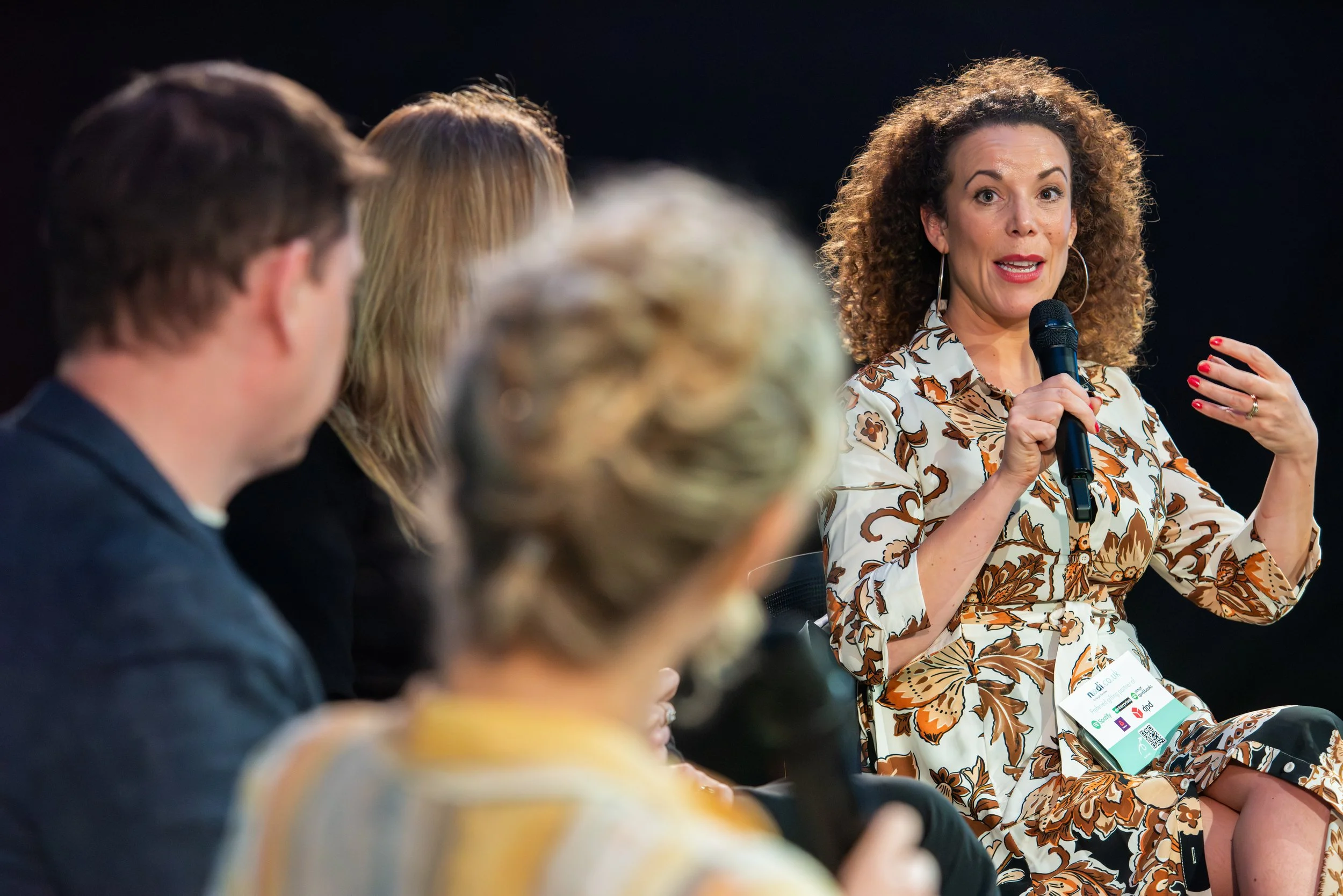 A woman with curly hair wearing a floral patterned dress and hoop earrings holding a microphone and speaking, while sitting on a panel with other blurred individuals in the foreground.