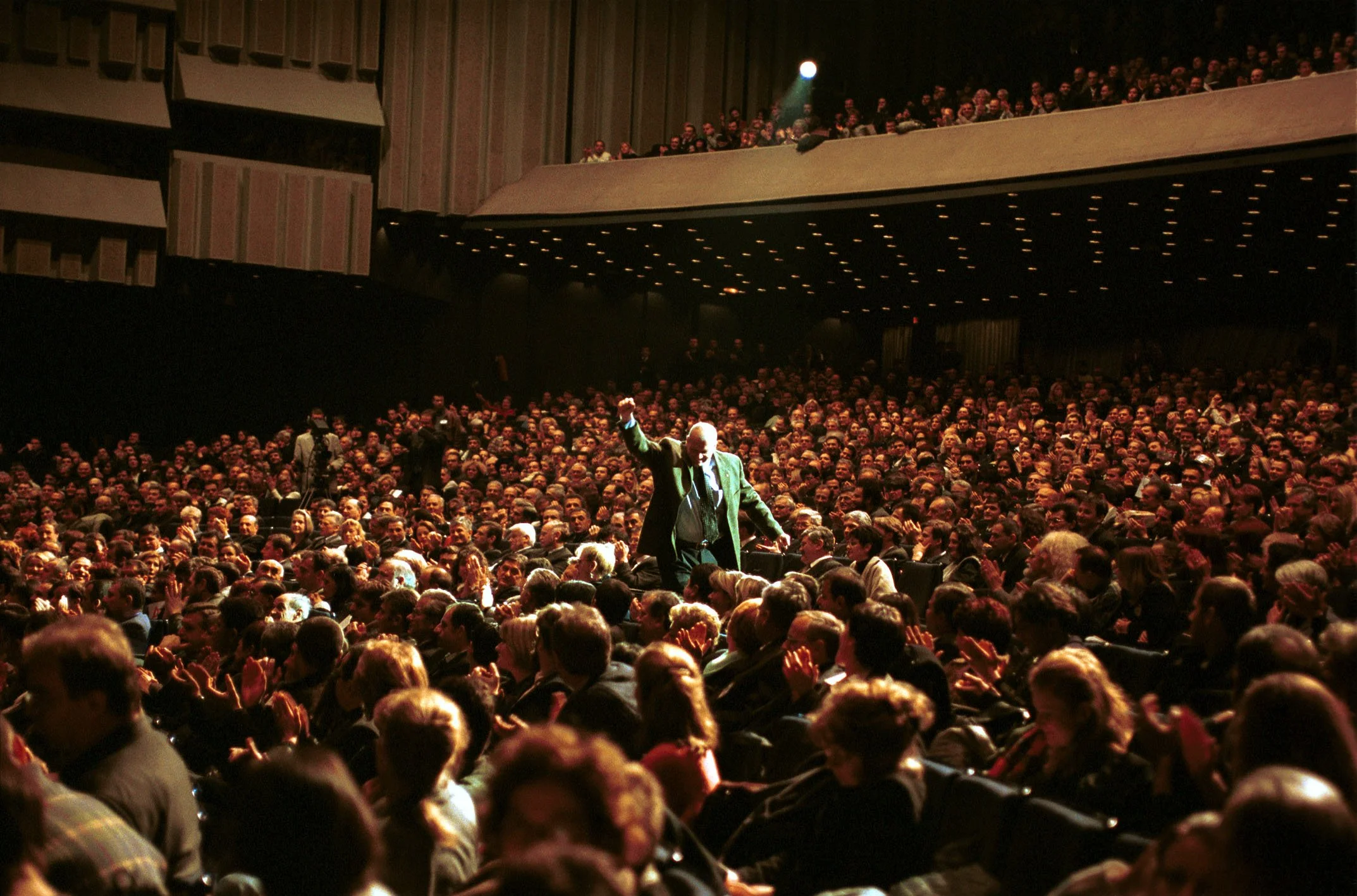 A large theater filled with an audience, with a man in a suit standing on stage in front of the crowd, raising his hand.