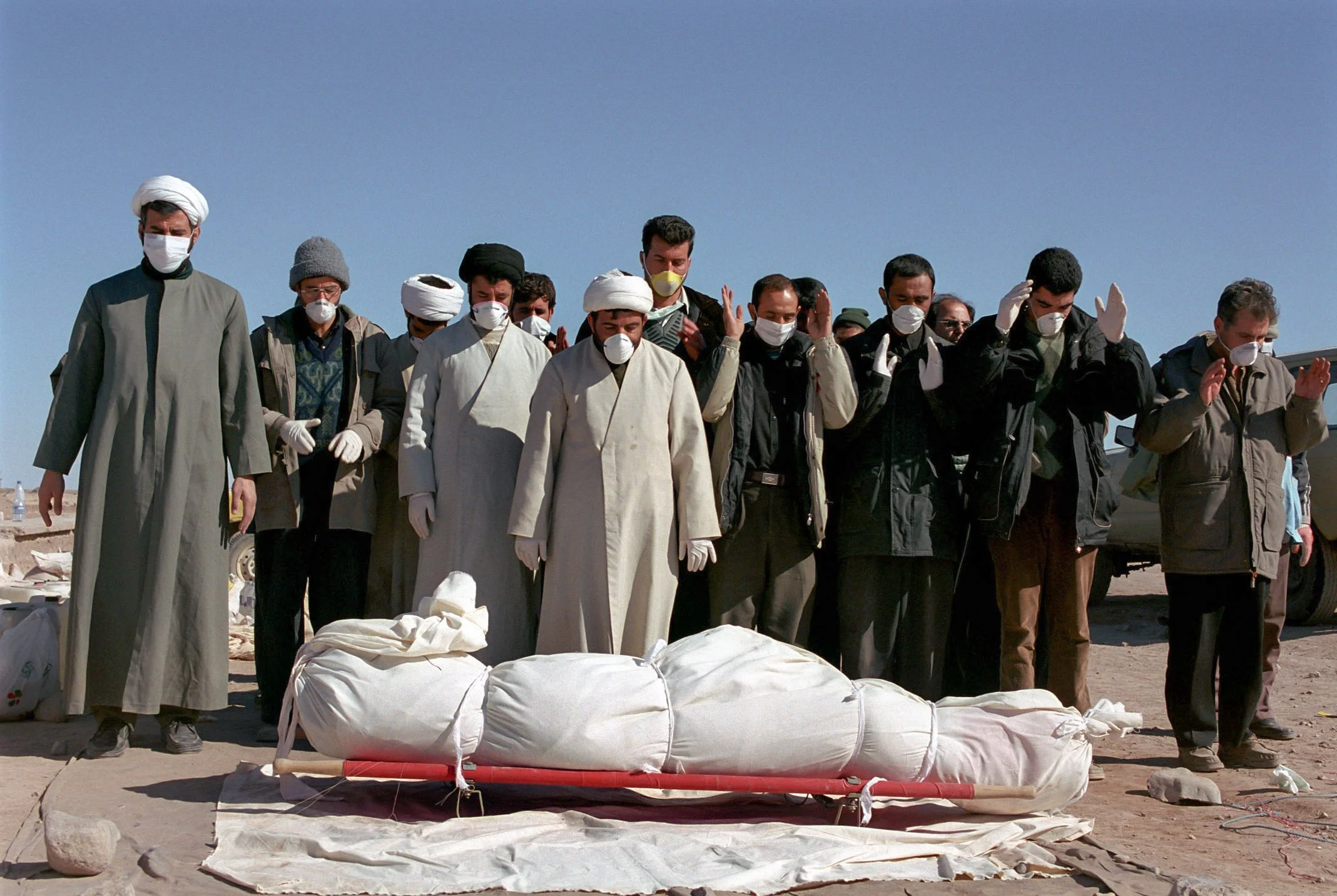 A group of people wearing face masks standing outdoors in a prayer position with hands raised or clasped. They are gathered around a large, wrapped object lying on a cloth on the ground, which appears to be a body or a figure wrapped in white cloth. 