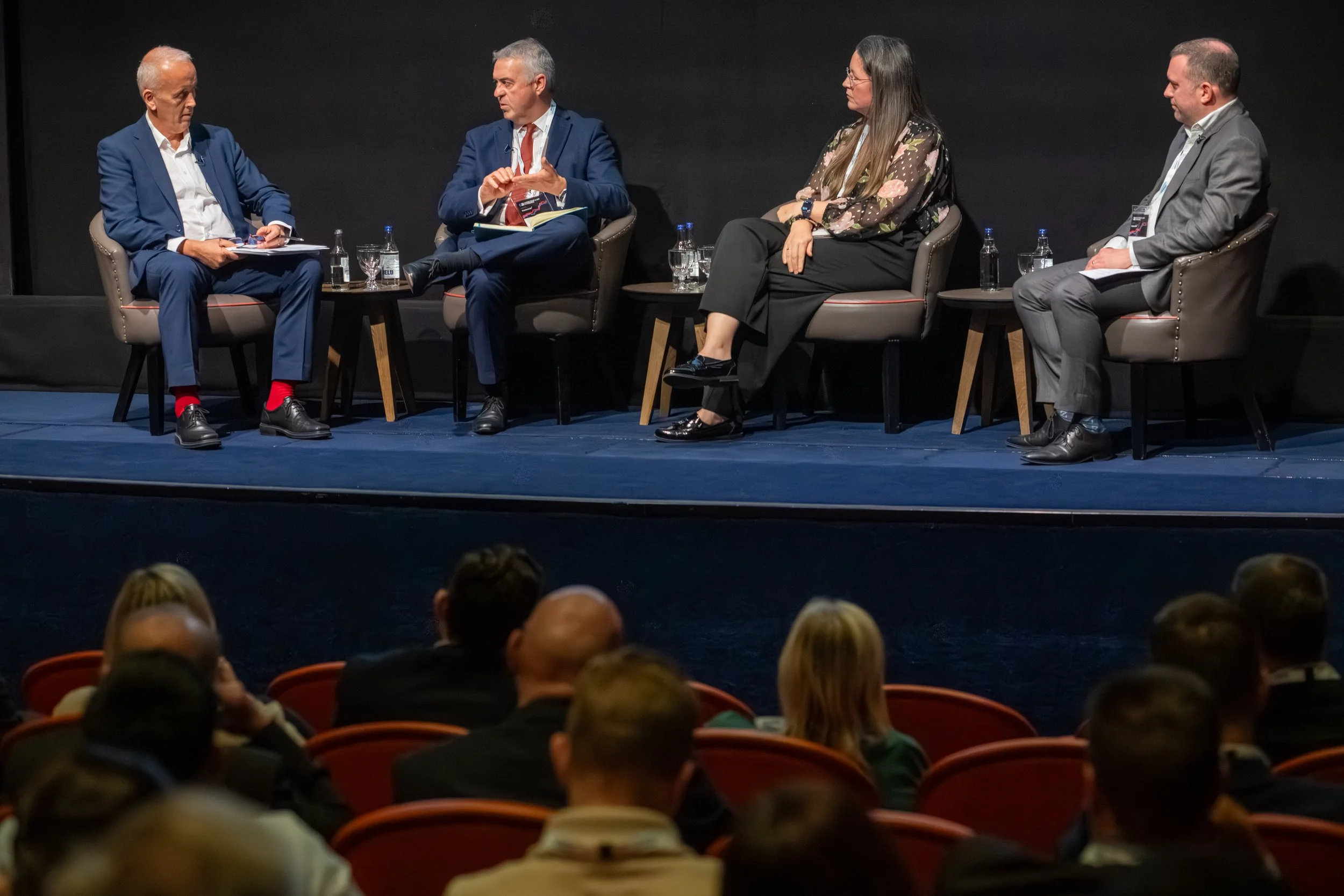 A panel discussion with four people seated on stage, including three men and one woman, with an audience in front of them.