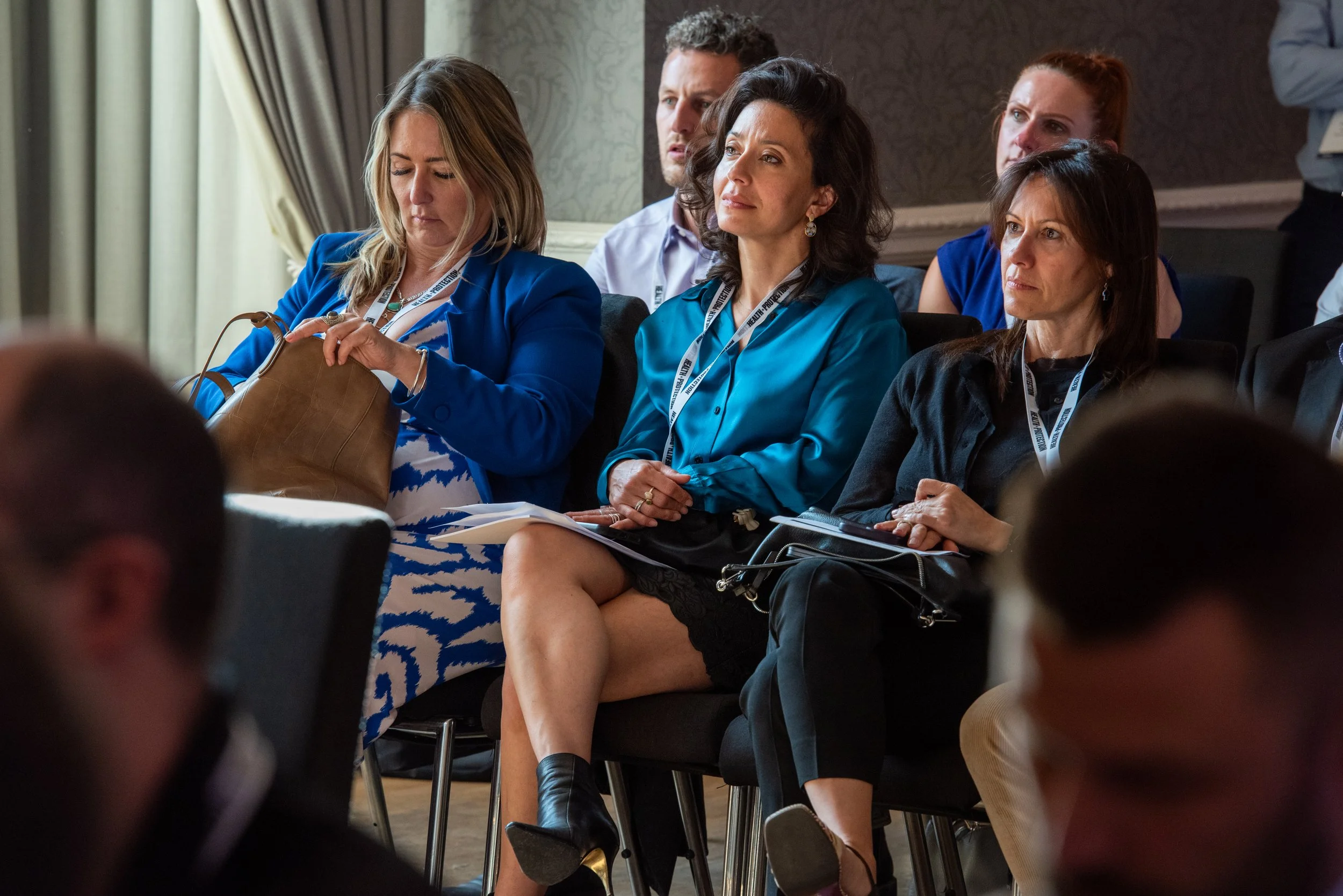 Group of professionals attending a conference or seminar, sitting in chairs, some taking notes, listening attentively.