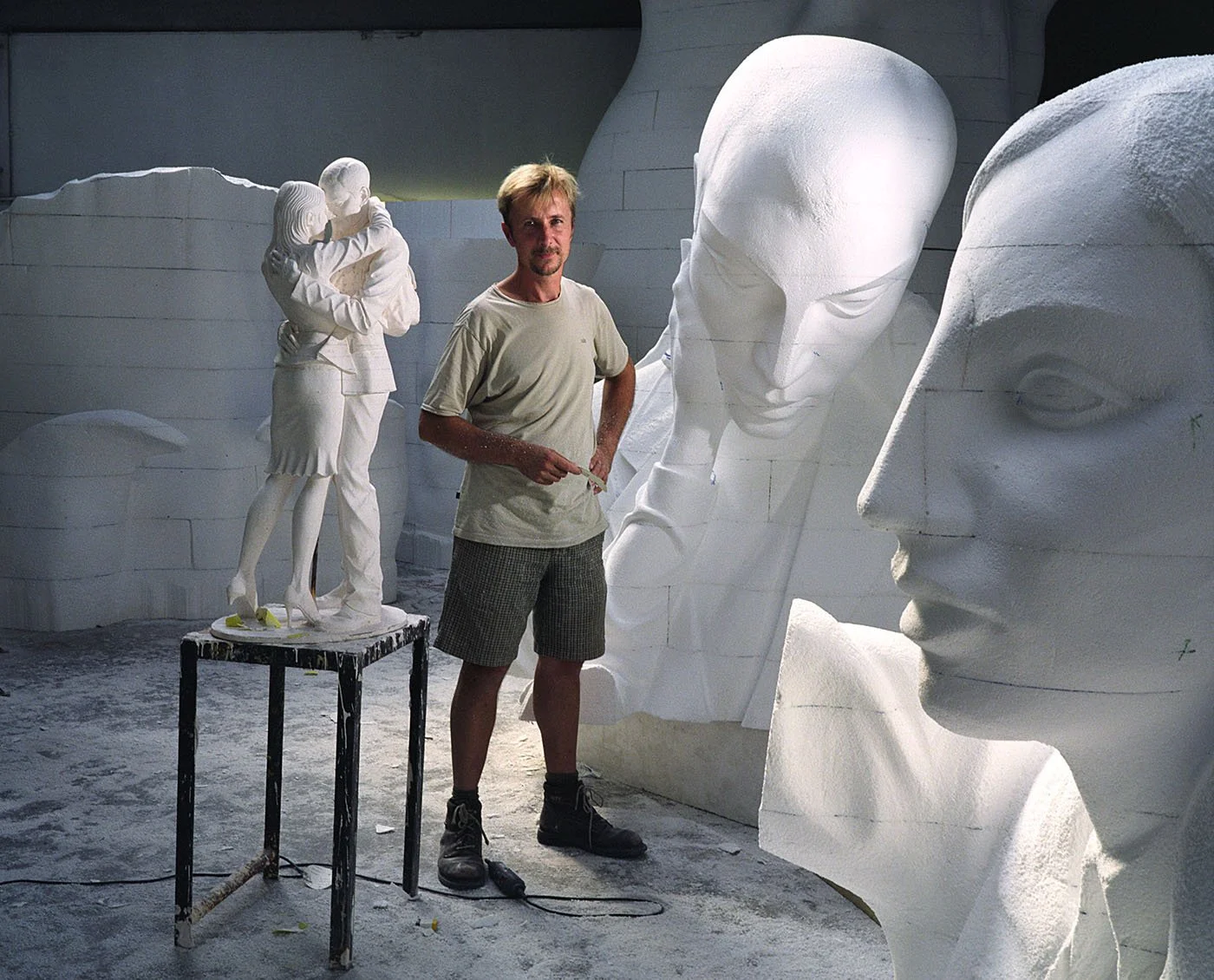 An artist working on sculptures of human faces and a hugging couple, all made of white material, in a studio with a concrete floor.