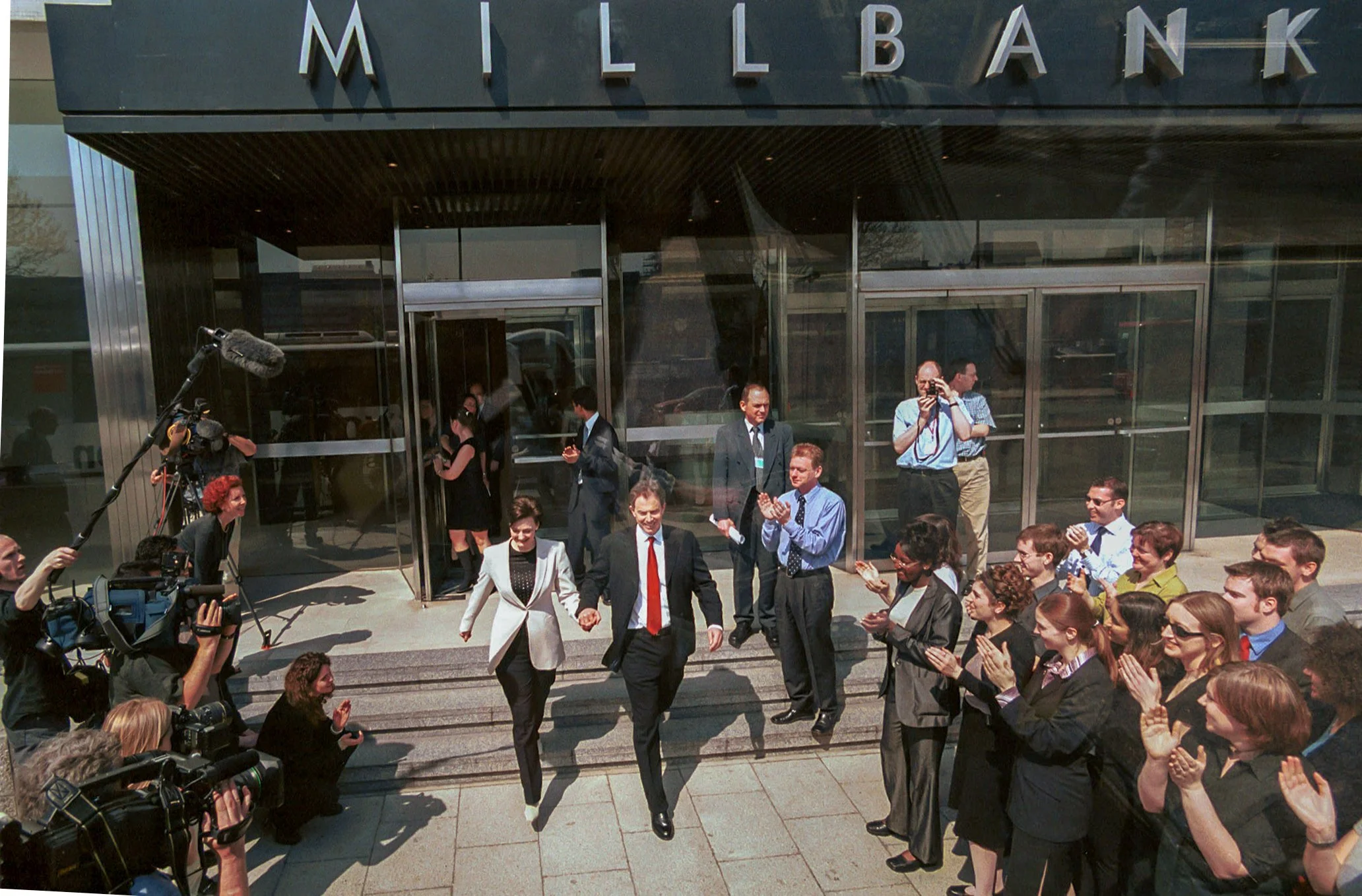 Prime Minister Tony Blair and his wife, Cheri, walking down the steps, flanked by a msmall crod on either side, outside of a building with a sign reading 'Millbank.' The crowd is clapping, with cameras and media personnel capturing the moment.