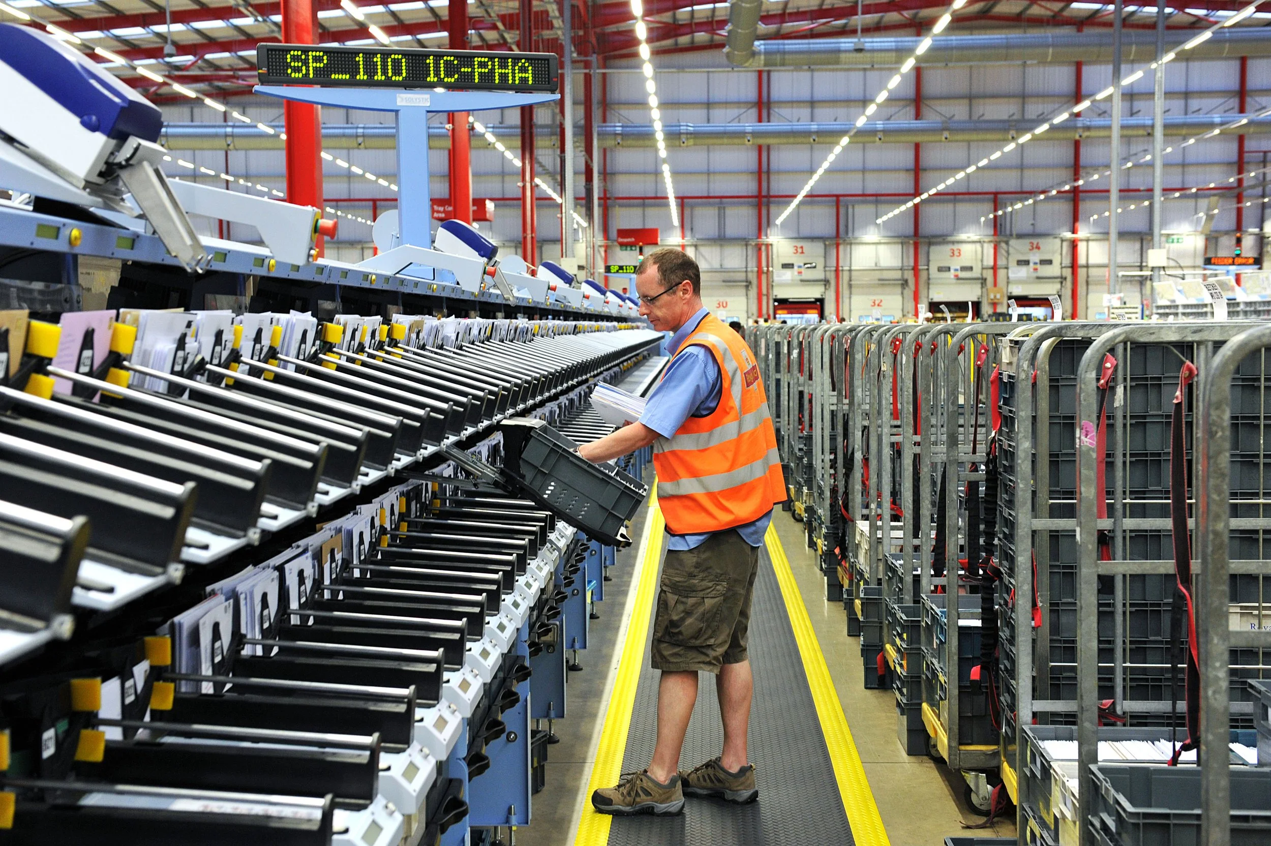 A warehouse worker in a blue shirt, orange hi-vis safety vest, and shorts is inspecting packages on a conveyor belt in a large industrial warehouse.