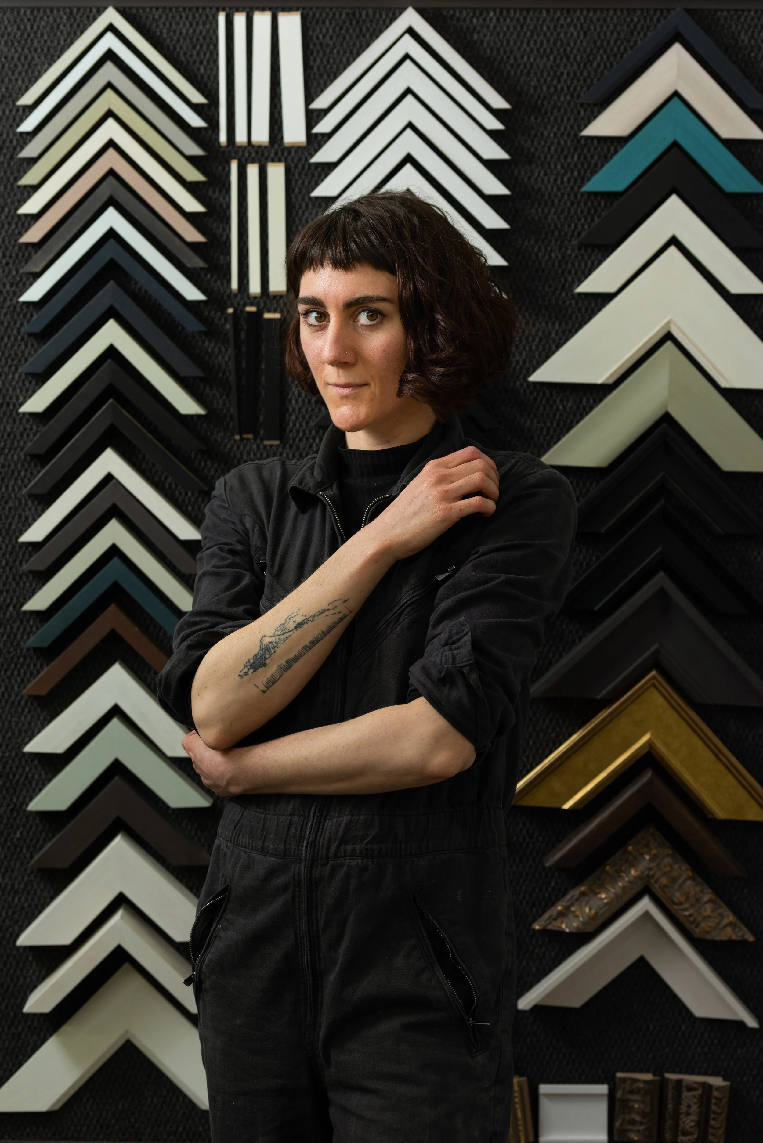 A woman with short dark hair, wearing a black jumpsuit, standing with arms crossed, in front of a display of various picture frame corners on a wall.