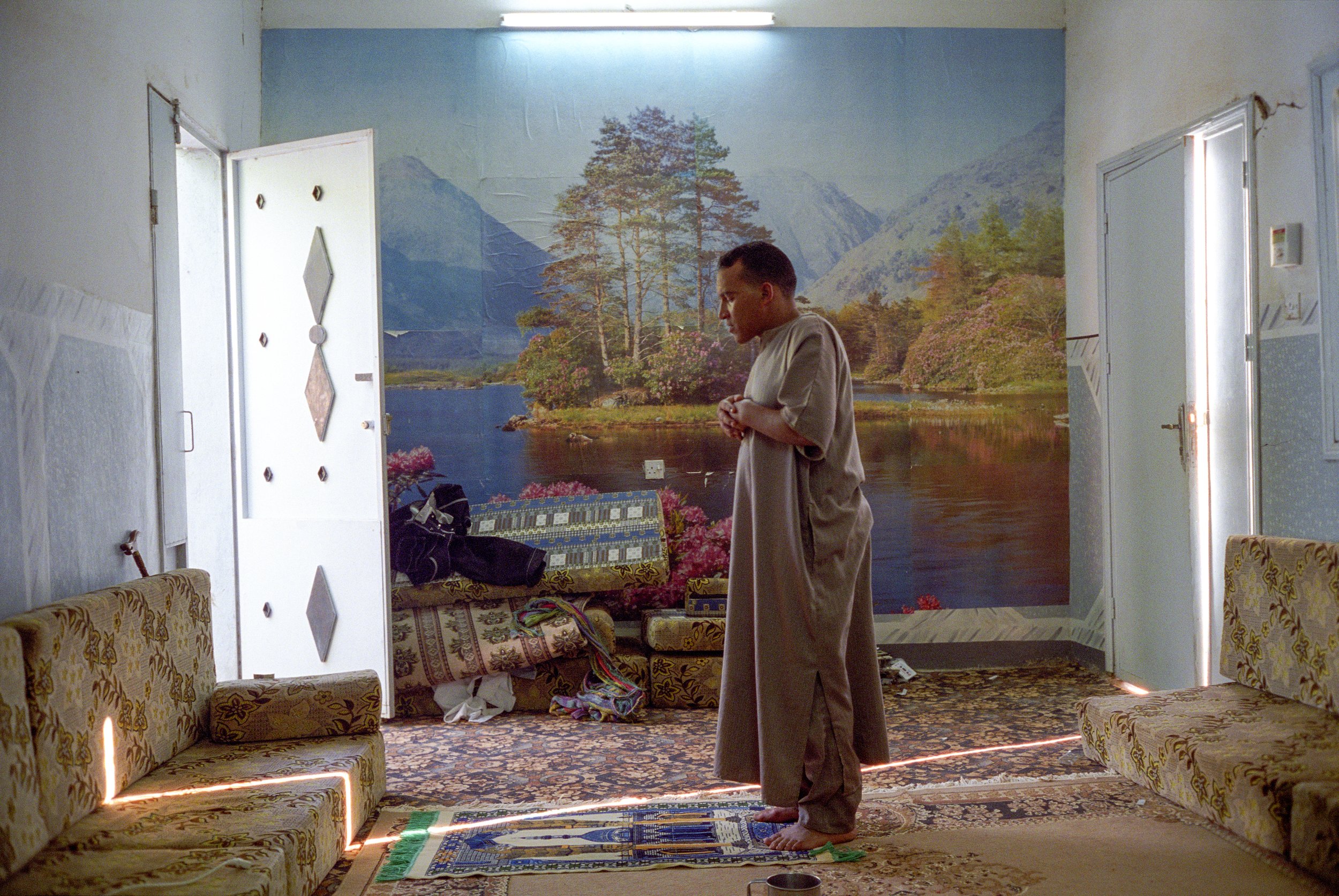 A praying young man standing in a room with a scenic mountain and lake mural on the back wall, with his hands clasped, dressed in loose clothing, surrounded by couches and a carpet.