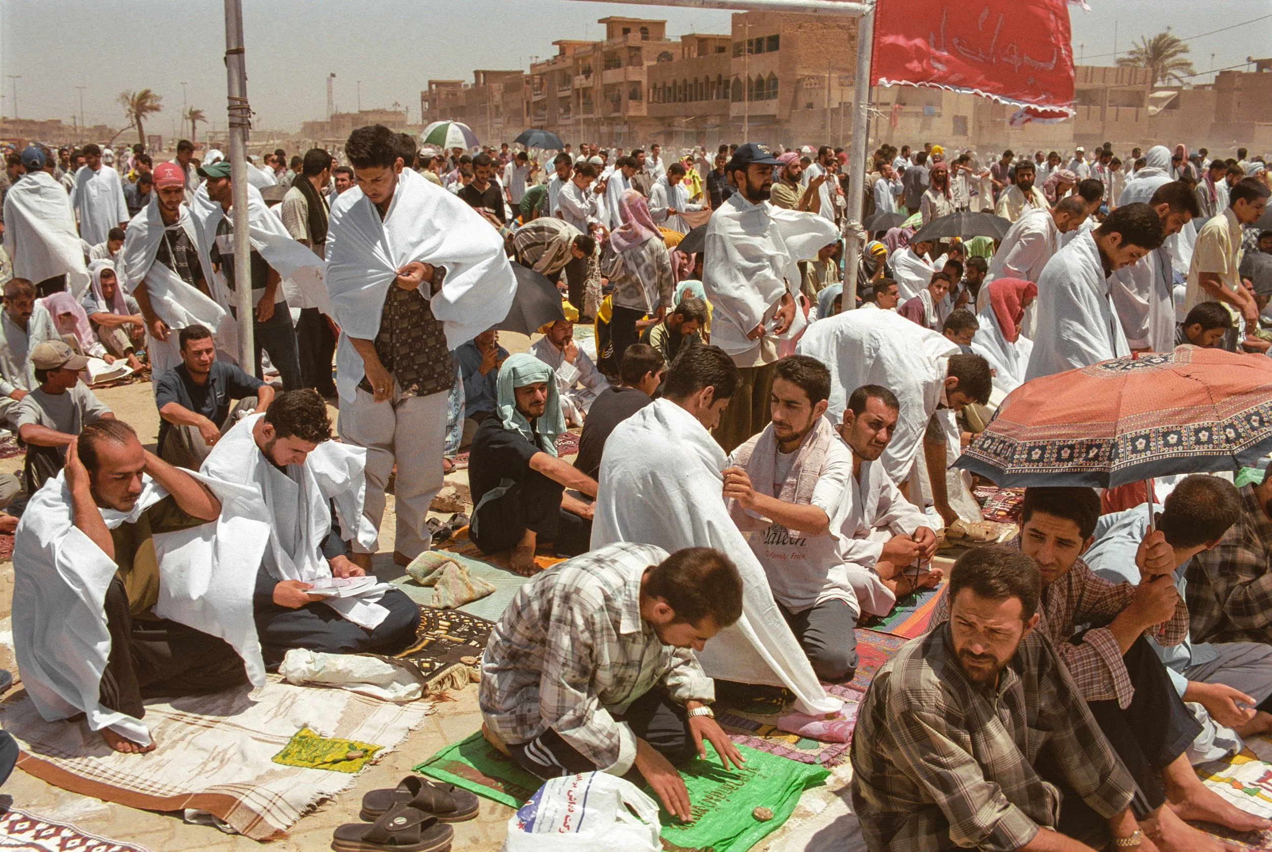 Crowd of men praying outdoors on prayer mats amid a dusty, sunny day with buildings and palm trees in the background.