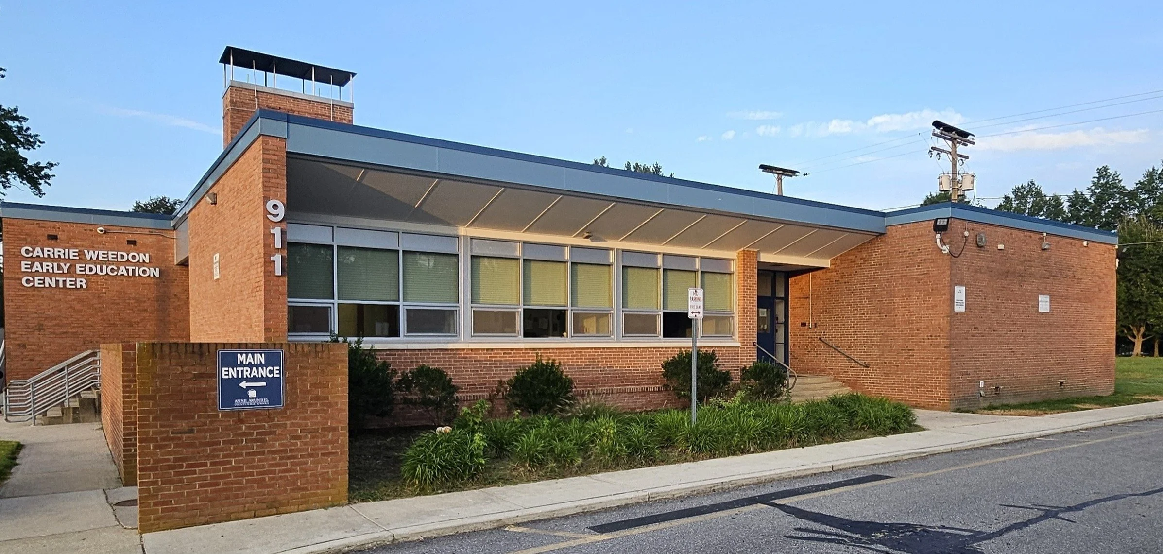 Exterior view of the Carrie Weedon Early Education Center, a brick building with a blue roof and large windows, with signs indicating the main entrance and street number 911.