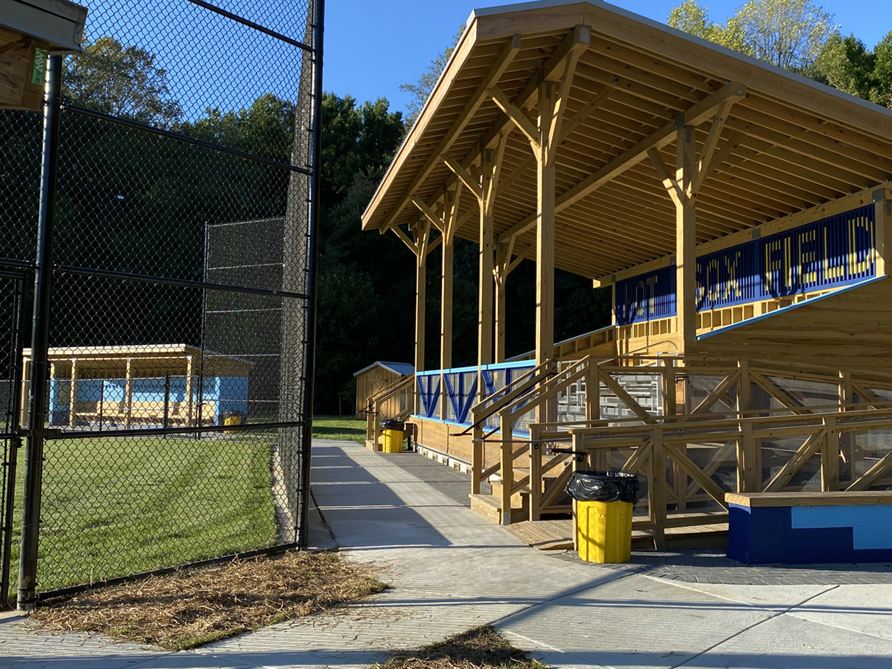 Galesville Hot Sox baseball field dugout with wooden supports and a blue sign, next to a chain-link fence surrounding the field, with grassy areas and trees in the background.