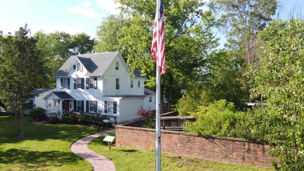 A white house with a black roof surrounded by green trees and a manicured lawn, with an American flag on a flagpole and a curved brick pathway leading up to it.