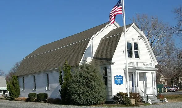 Galesville Memorial Hall with American flag, blue sign, and small front staircase, surrounded by trees and shrubs.
