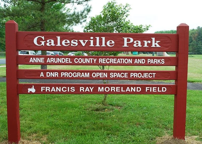 Sign at Galesville Park with red wood frame, displaying park name and information about Anne Arundel County Recreation and Parks, a DNR open space project, and Francis Ray Moreland Field, with trees and grass in the background.