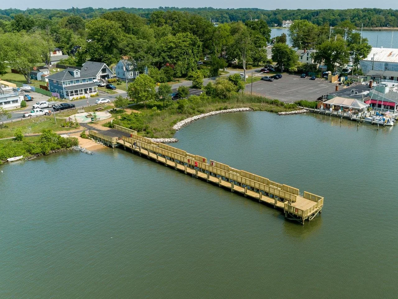 Aerial view of a Galesville pier extending into a body of water, with a small sandy beach area and trees along the shore, residential houses in the background, and a parking lot near the water.