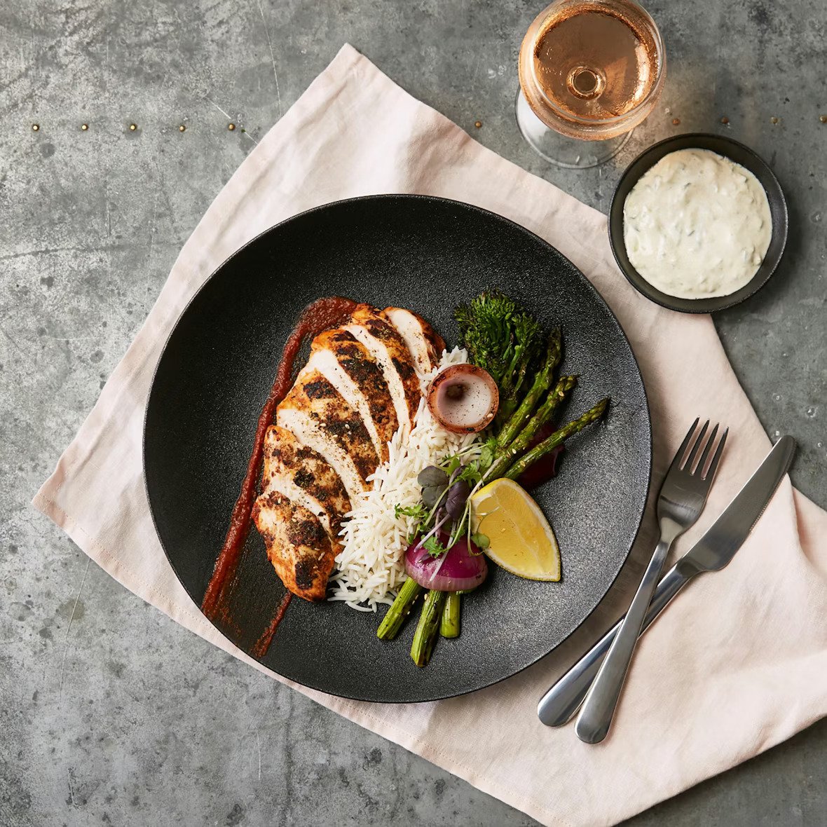 Grilled chicken breast with barbecue sauce on a black plate, served with rice, grilled asparagus, broccoli, radish, lemon wedge, and a slice of onion. A glass of rosé wine and a small bowl of creamy dipping sauce are on the side.