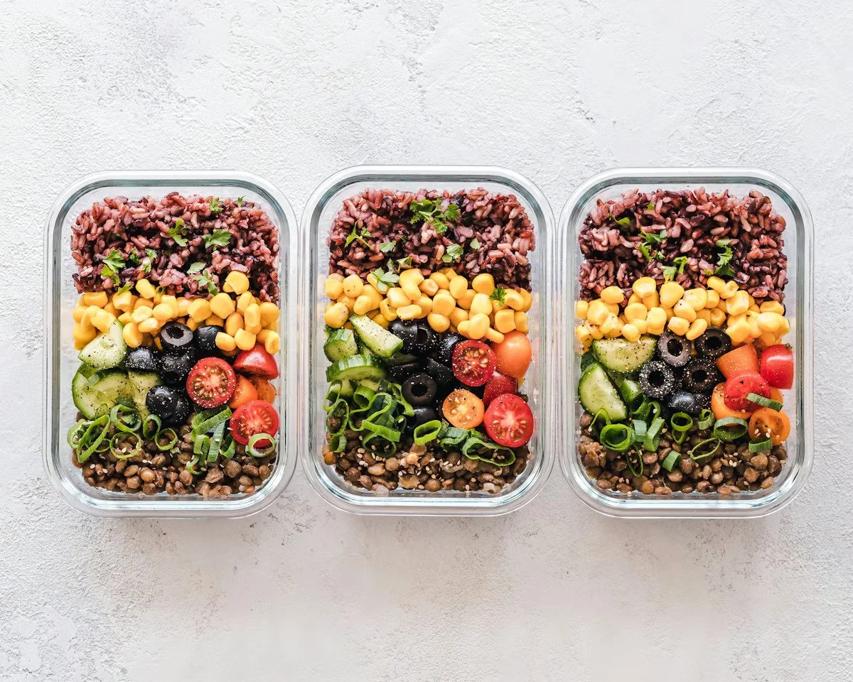 Three glass meal prep containers filled with layered salads, including black beans, cherry tomatoes, cucumbers, chickpeas, lentils, green onions, and mixed grains on a white textured surface.