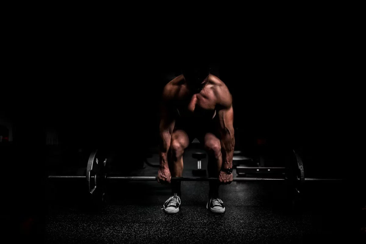 A muscular man in black athletic wear performing a deadlift exercise in a dark gym, with a loaded barbell on the floor.