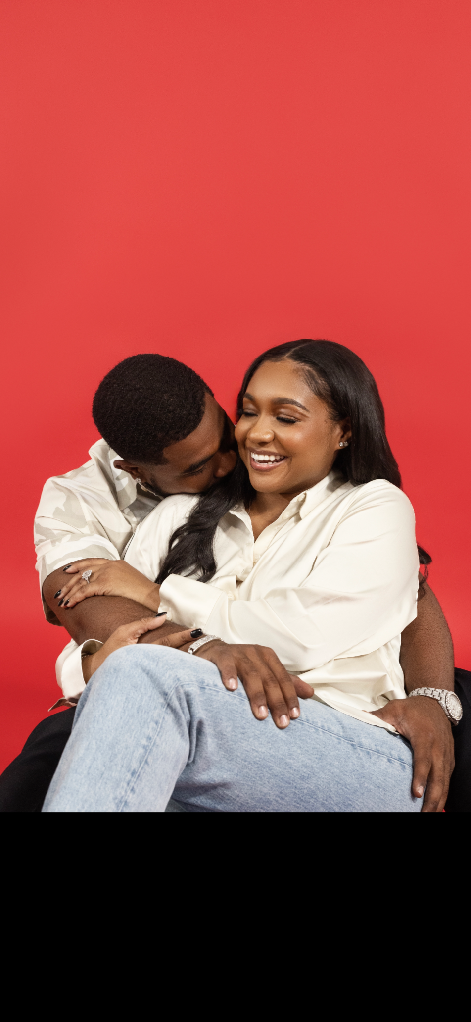 A happy African American couple sitting close together against a red background, embracing and smiling in a candid, intimate moment.