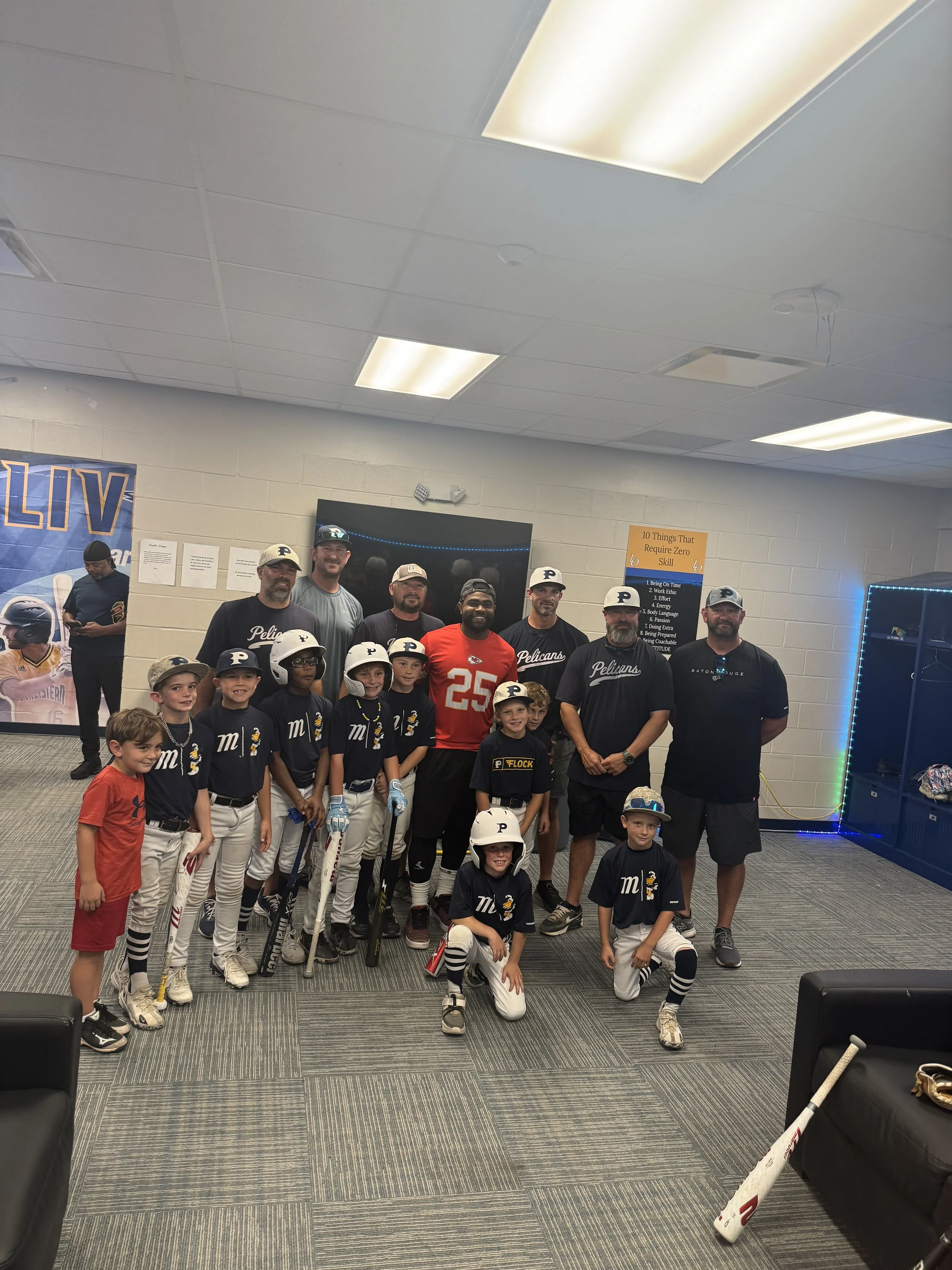 A group of young baseball players in uniform and their coaches posing for a photo indoors. The kids are wearing black jerseys with a letter M, white pants, and some have helmets. Some coaches are wearing black shirts, and one is in a red jersey with the number 25. The group is standing on a patterned carpet with a dark TV behind them. The room has a white brick wall, a poster, and some sports memorabilia.