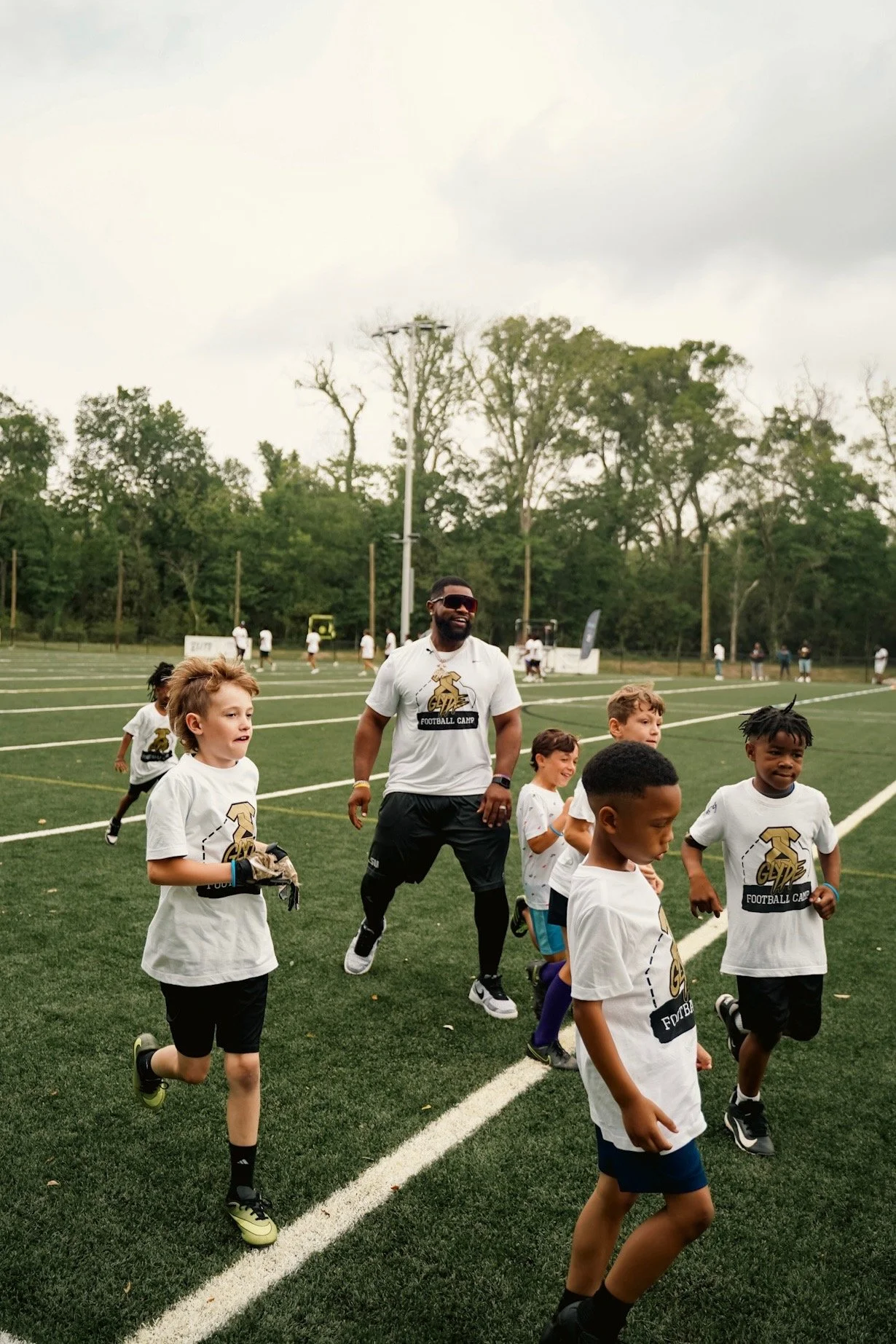 A group of children and a coach running on a football field during a sports camp. The children are wearing white T-shirts and athletic shorts, while the coach is dressed in sportswear.