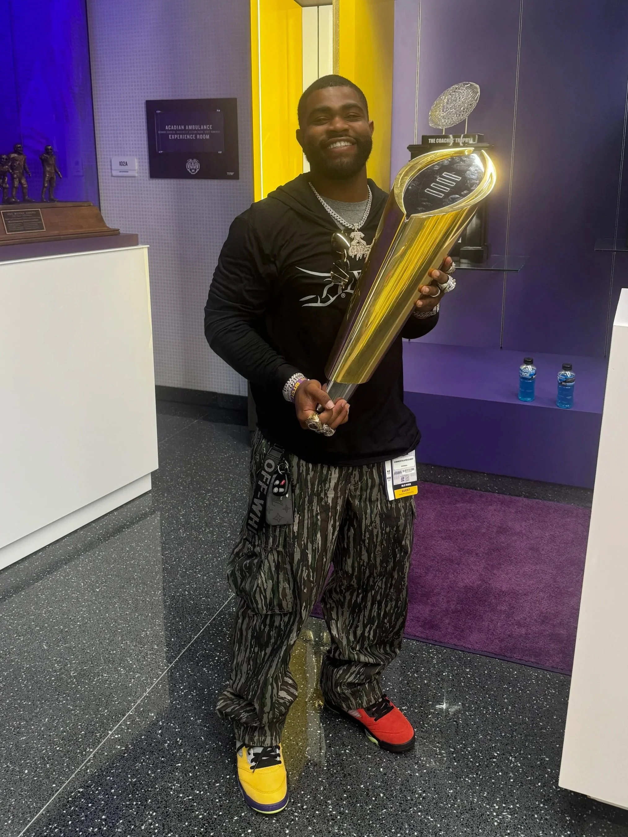 A man smiling and holding a large golden trophy in an exhibit room with purple and yellow walls. He is wearing a black hoodie, patterned cargo pants, and colorful sneakers, with jewelry and sunglasses hanging from his hoodie.